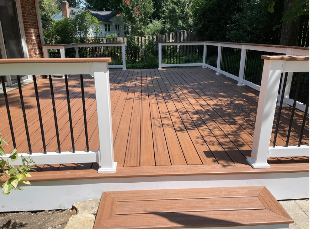 Newly built wooden deck with white and black railing, outside a house with trees and greenery in the background.