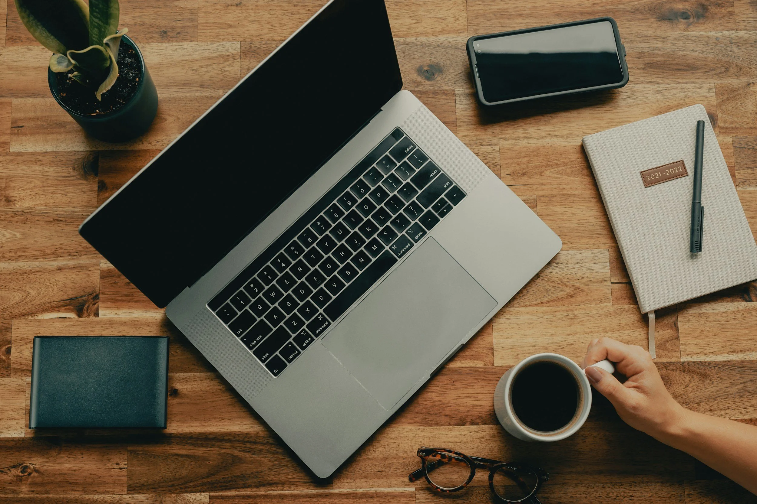 A wooden desk with a silver laptop, a potted plant, a black notebook, a smartphone, a closed notebook with a leather patch labeled 2021-2022, a pen, a pair of glasses, and a person's hand holding a coffee mug.