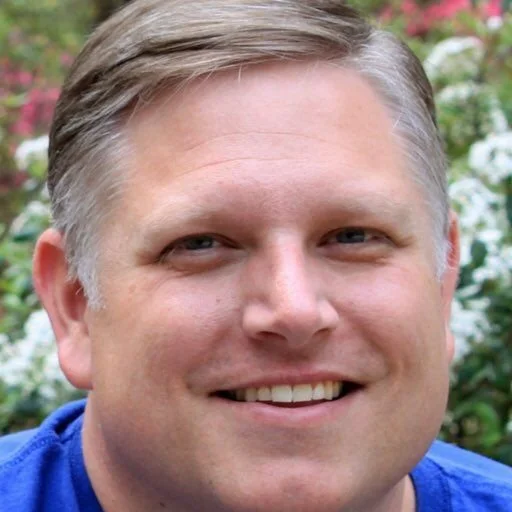 Close-up photo of a smiling man with short, light brown hair and blue eyes, outdoors with blurred greenery and flowers in the background.
