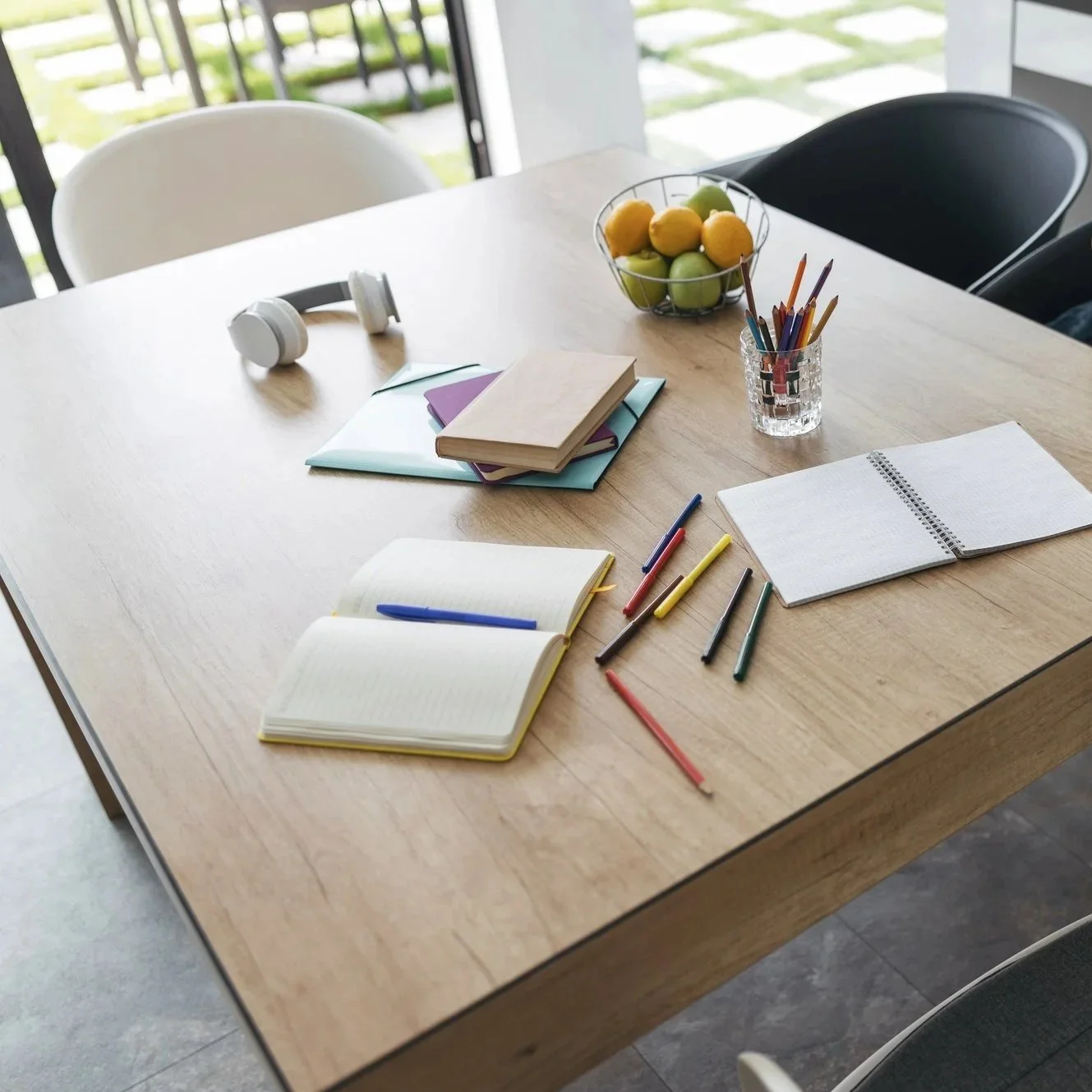 A light wooden table with notebooks, colored pens, a bowl of lemons and limes, a pair of white headphones, and two chairs on either side.