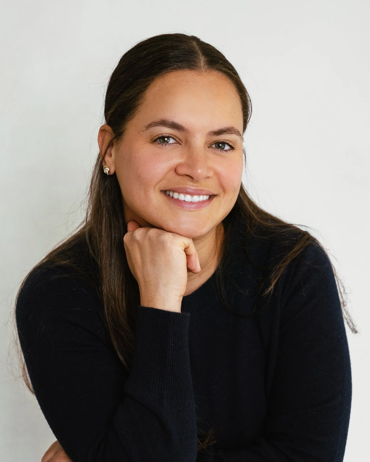 Close-up of a woman with long dark brown hair, wearing a black sweater and stud earrings, smiling with her chin resting on her hand in front of a plain white background.