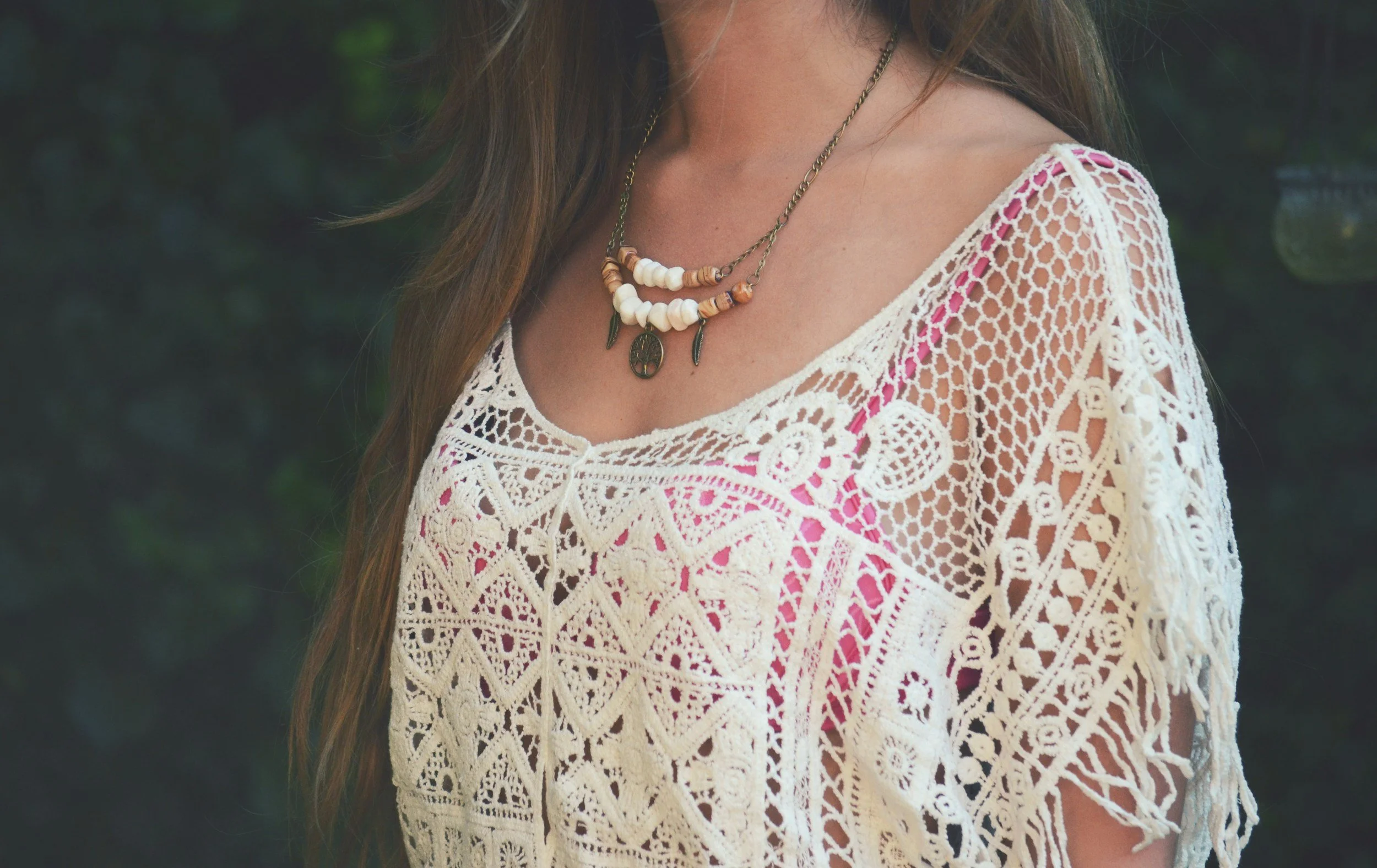 A woman wearing a white crochet top with pink accents, layered necklaces including white beads, wood beads, and pendants, with long brown hair.
