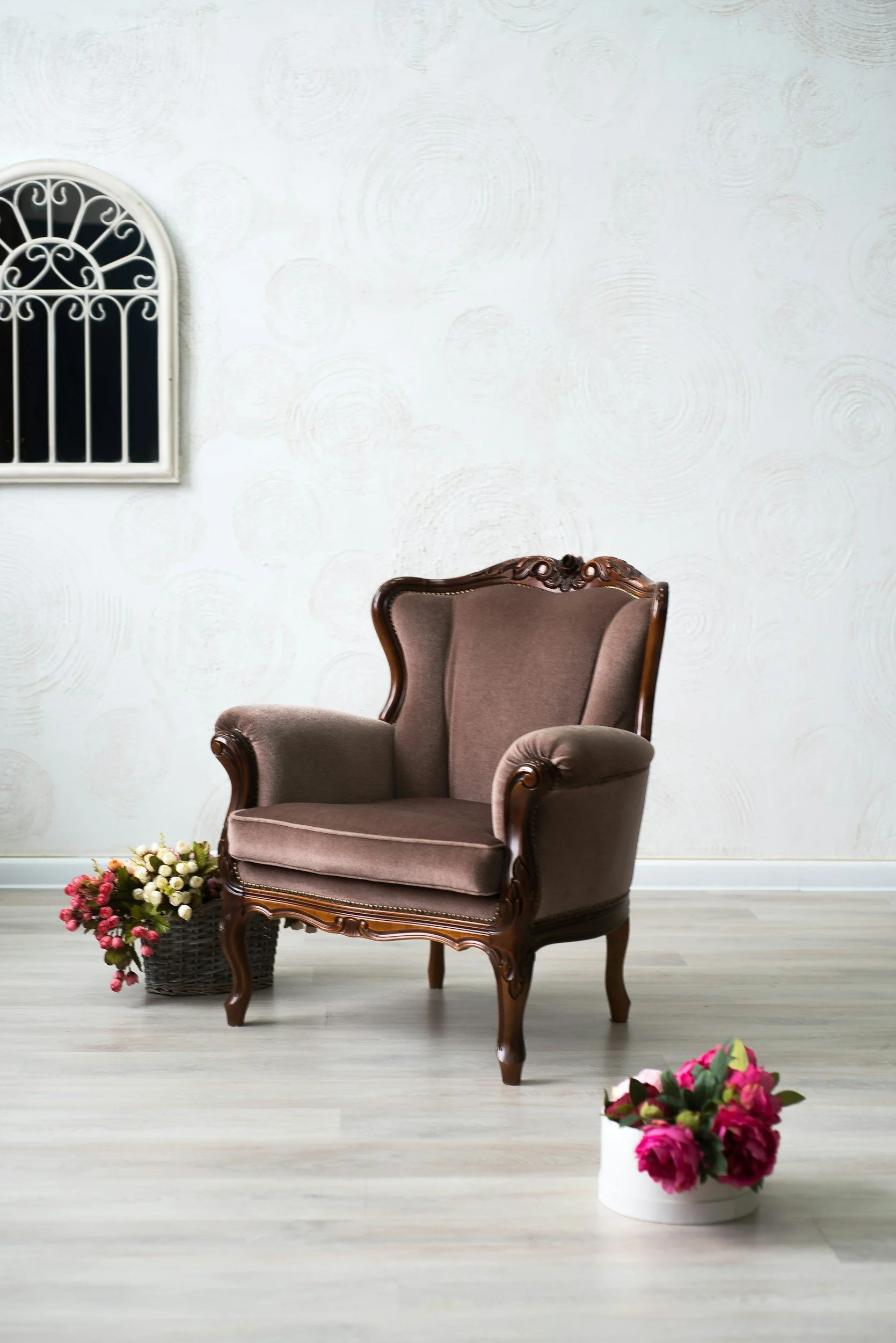 A vintage armchair with carved wooden details and brown upholstery, placed in a room with light-colored flooring and a white wall with textured circular patterns. There are two baskets of pink and white flowers, one on the left side and one in the foreground.