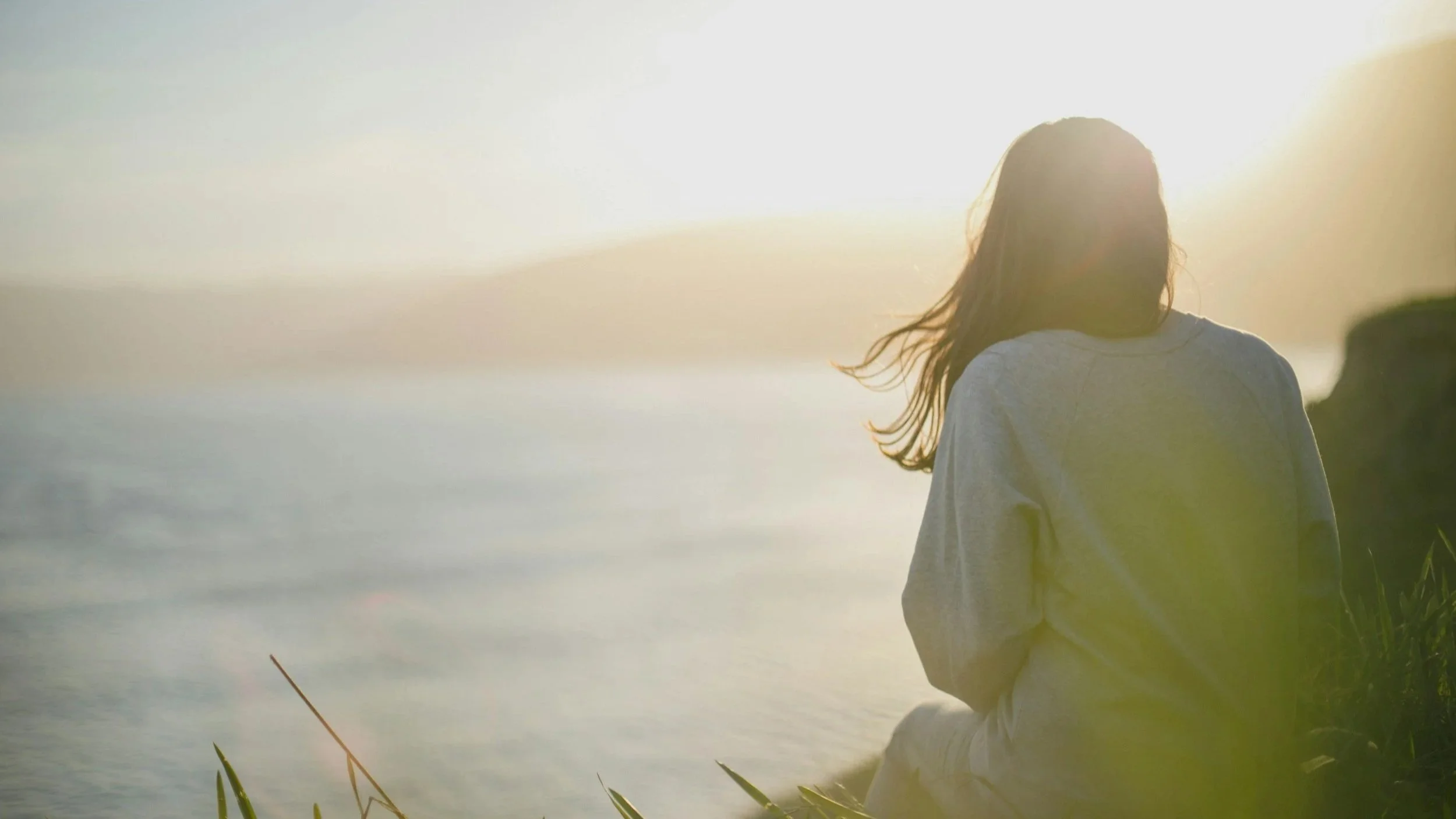 A woman stands near a body of water during sunrise or sunset, with her back to the camera, wearing a light-colored hoodie.