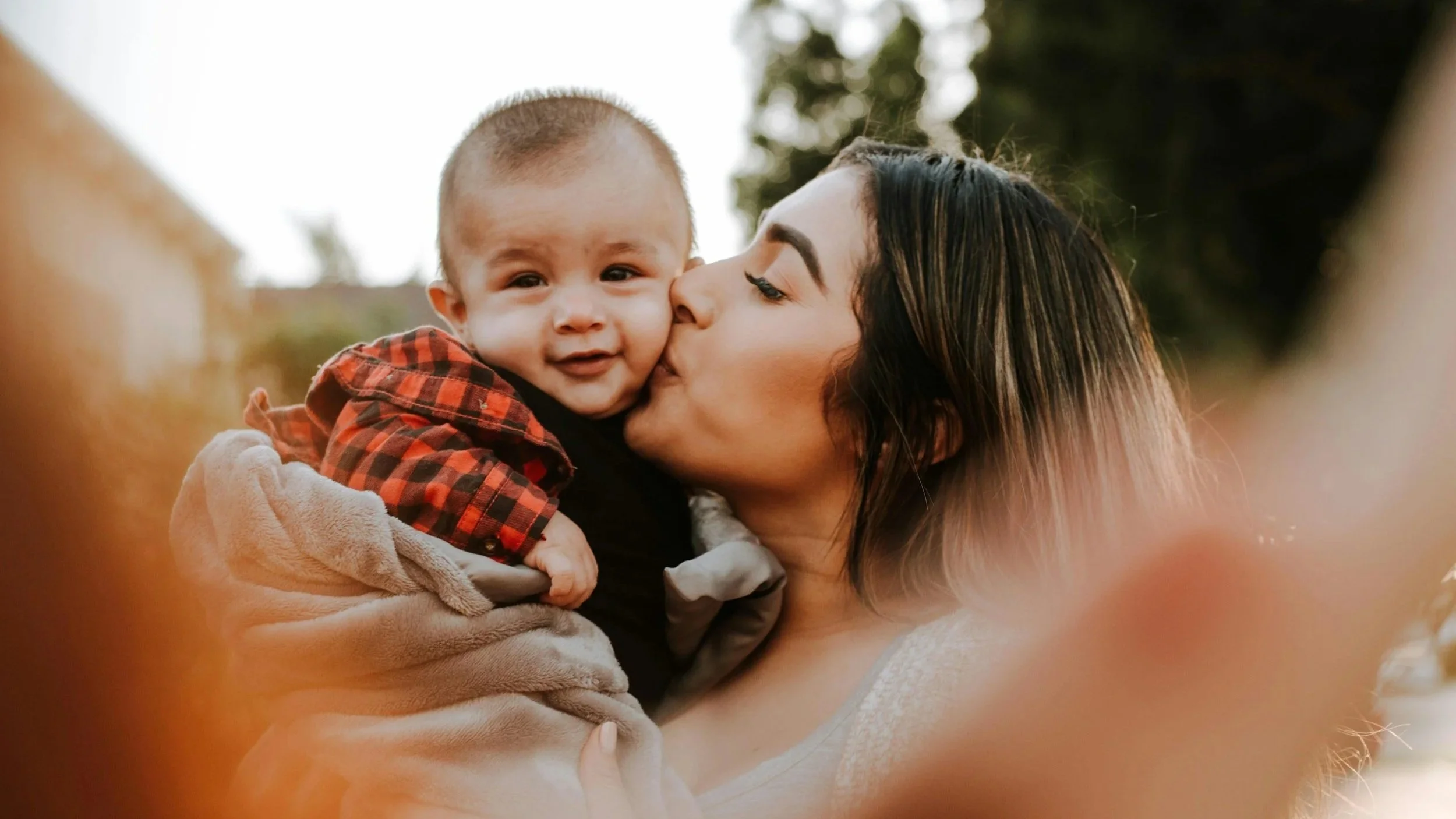 A woman with dark, long hair kissing a young boy on the cheek outdoors.