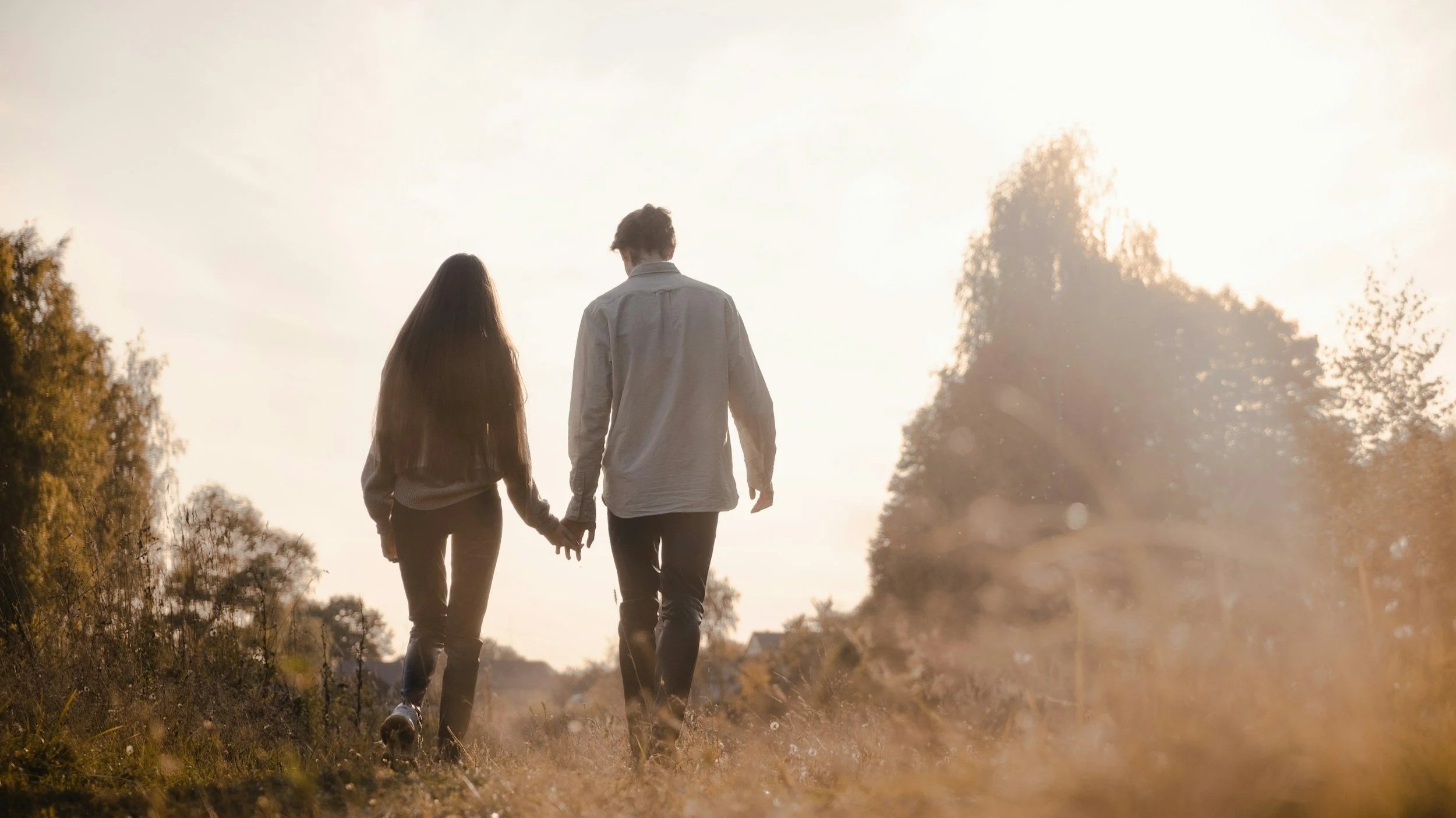 A couple walking hand-in-hand through a natural outdoor setting with trees, during sunset or sunrise.