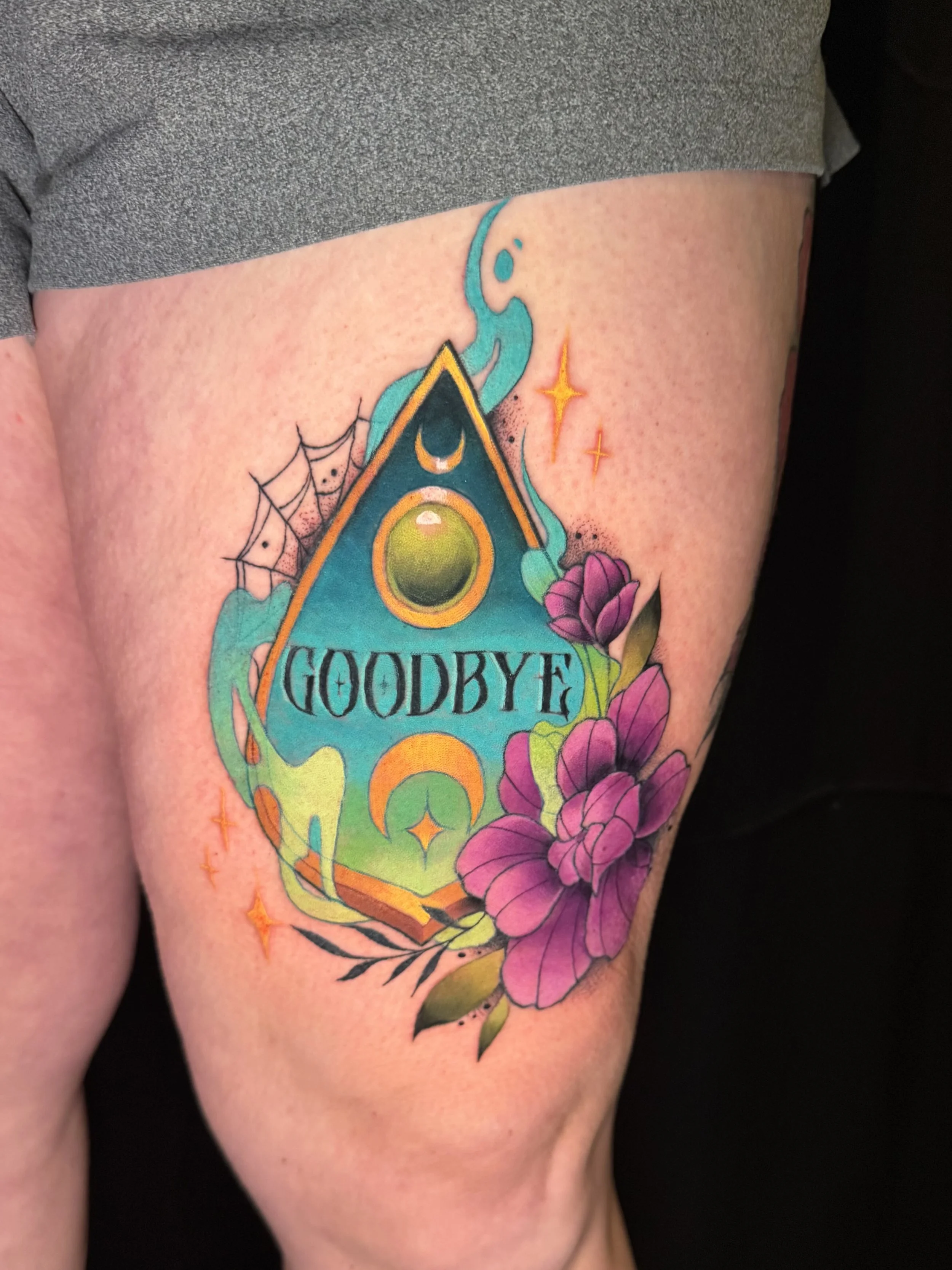 Colorful tattoo on the upper arm featuring a triangle with the word 'GOODBYE' inside, surrounded by flowers, a spider web, and small stars, with a blue snake-like figure emerging from the top.