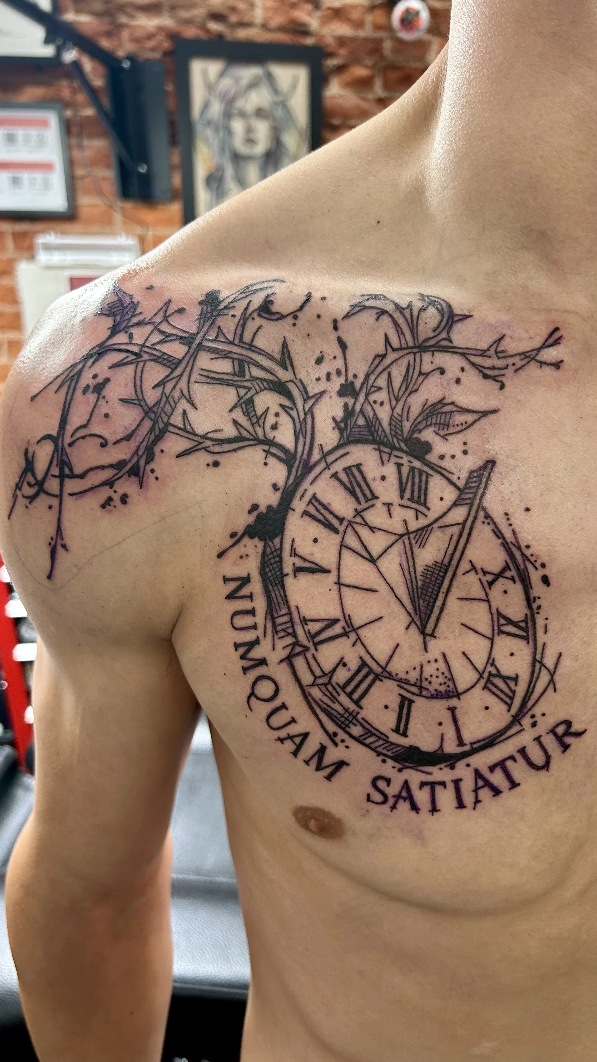 A person's upper chest and shoulder with a detailed black ink tattoo of a clock with Roman numerals, a branch with leaves, and Latin words. The background includes a brick wall and framed artwork.