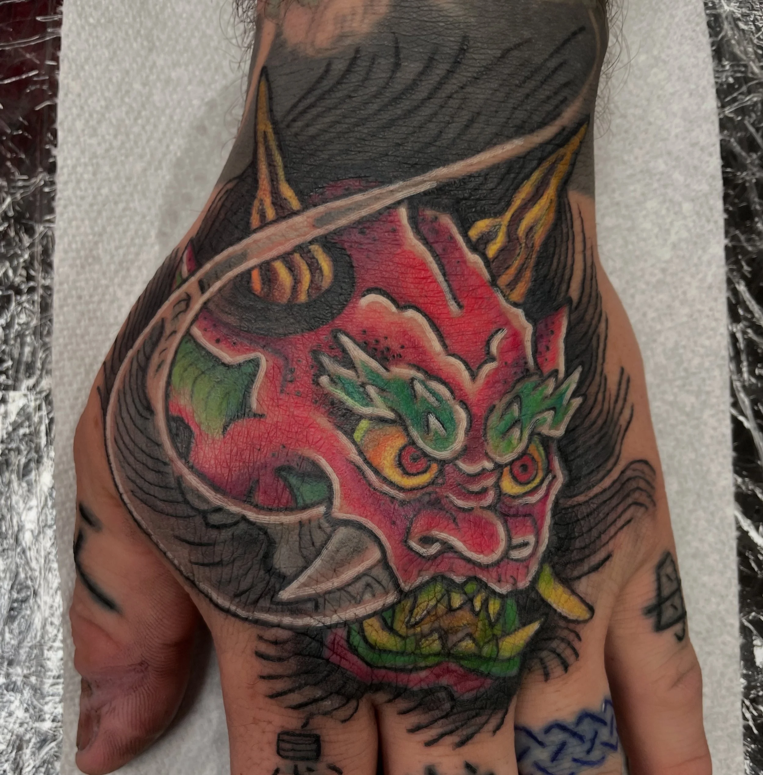 Close-up of a person's arm with tattooed skin showing colorful artwork featuring red, yellow, green, and black ink, along with shading and line work.