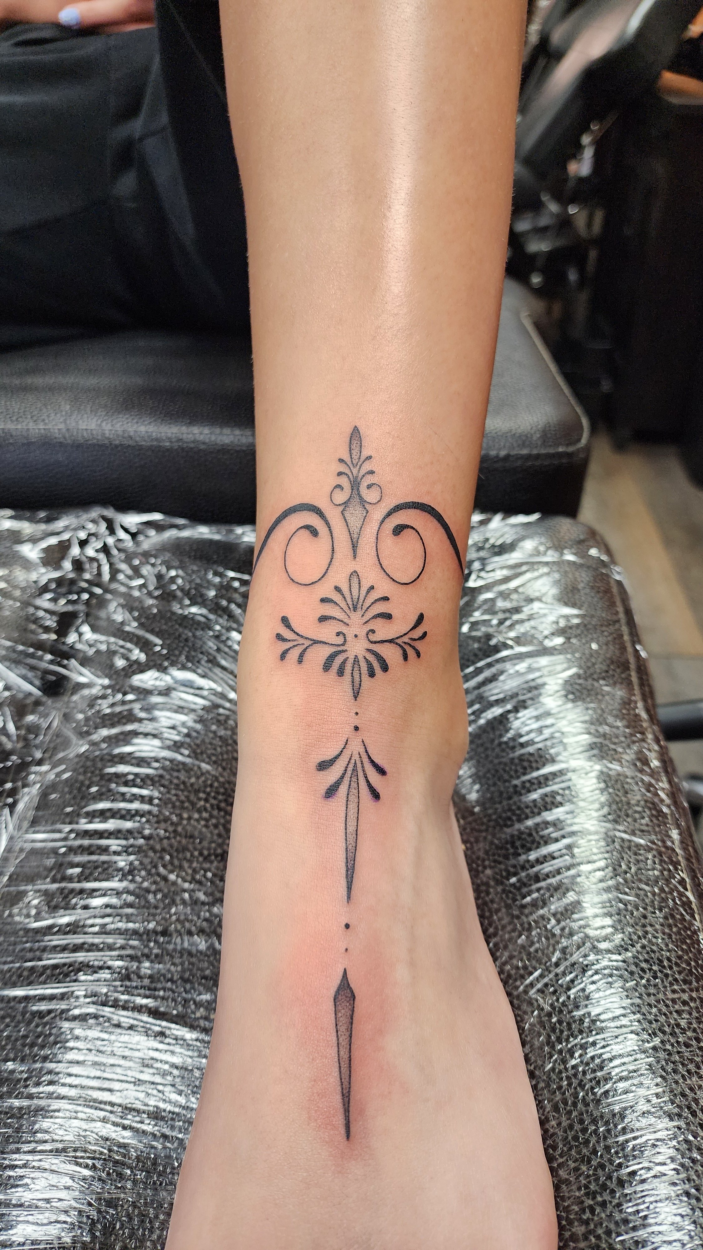 Tattoo on person's forearm featuring a symmetrical, black line design with flourishes and ornamental details.