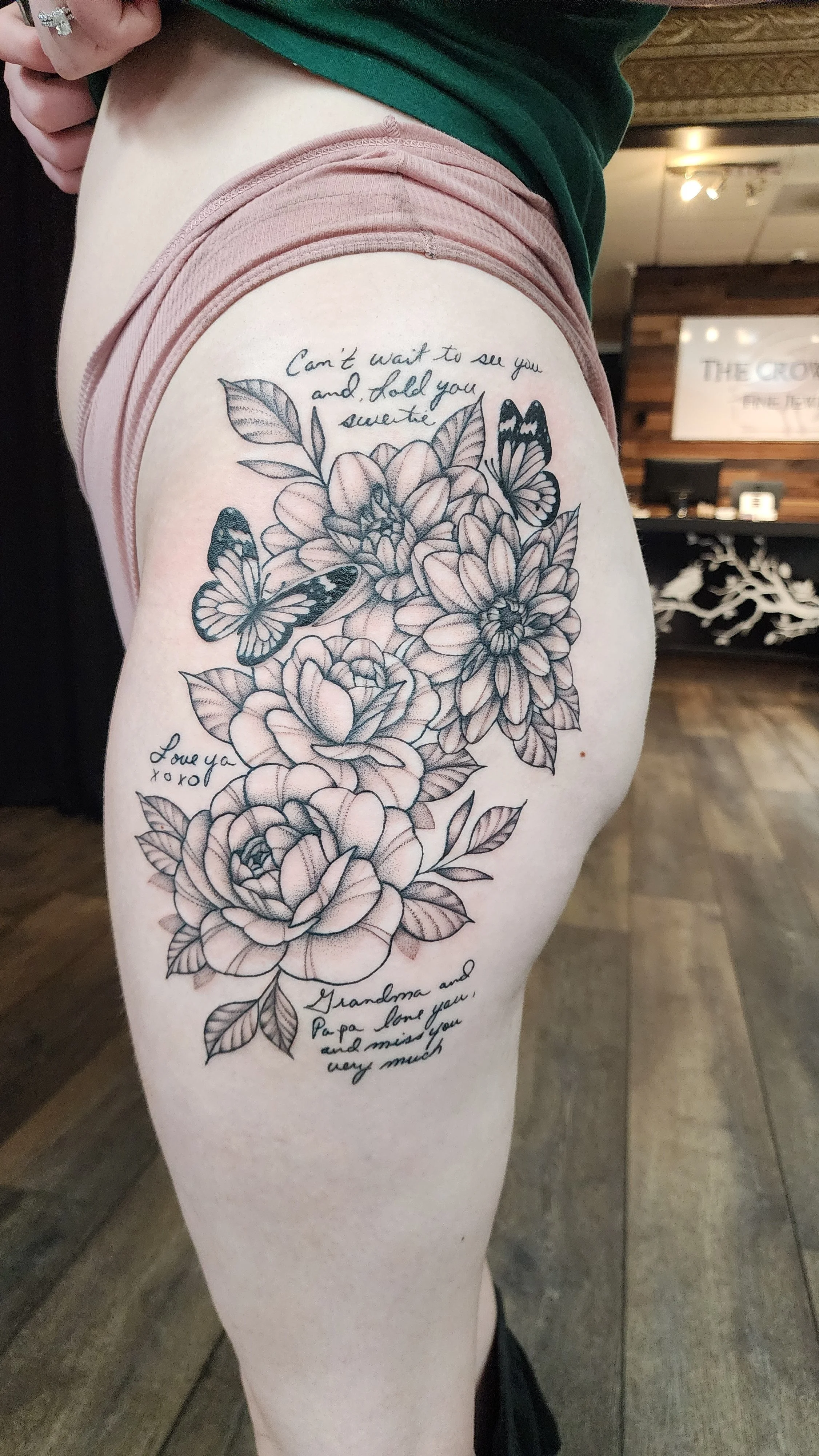 A tattoo of flowers and butterflies on a person's thigh with handwritten-style text. The visible part of the text reads, 'Can't wait to see you and hold you sweetie' and 'Love you xoxo.' There is also a message mentioning 'Grandma and Papa love you and miss you very much.'