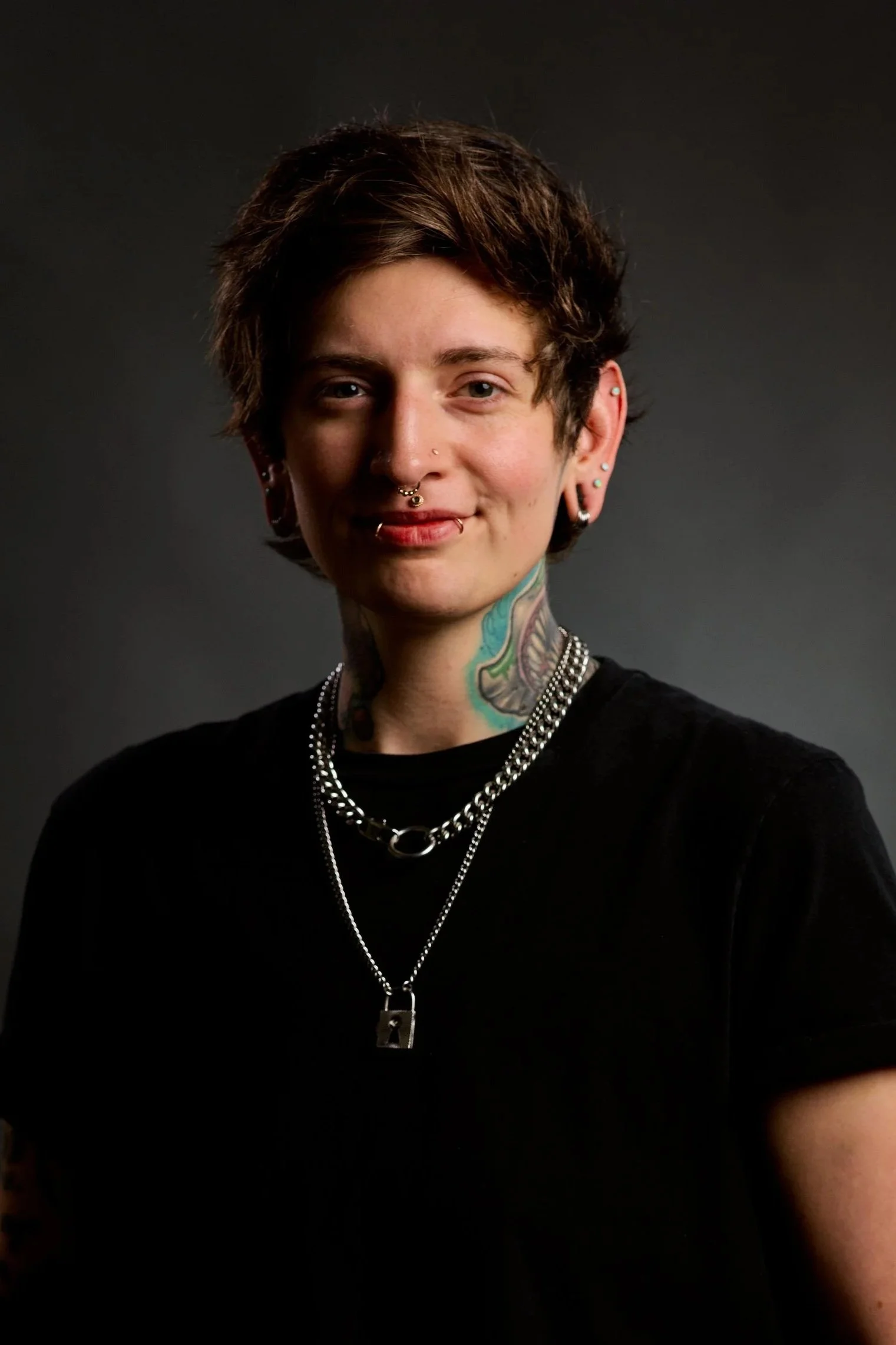 Young person with short, wavy brown hair, multiple facial piercings, and tattoos on their neck, wearing layered silver necklaces and a black shirt, against a dark background.