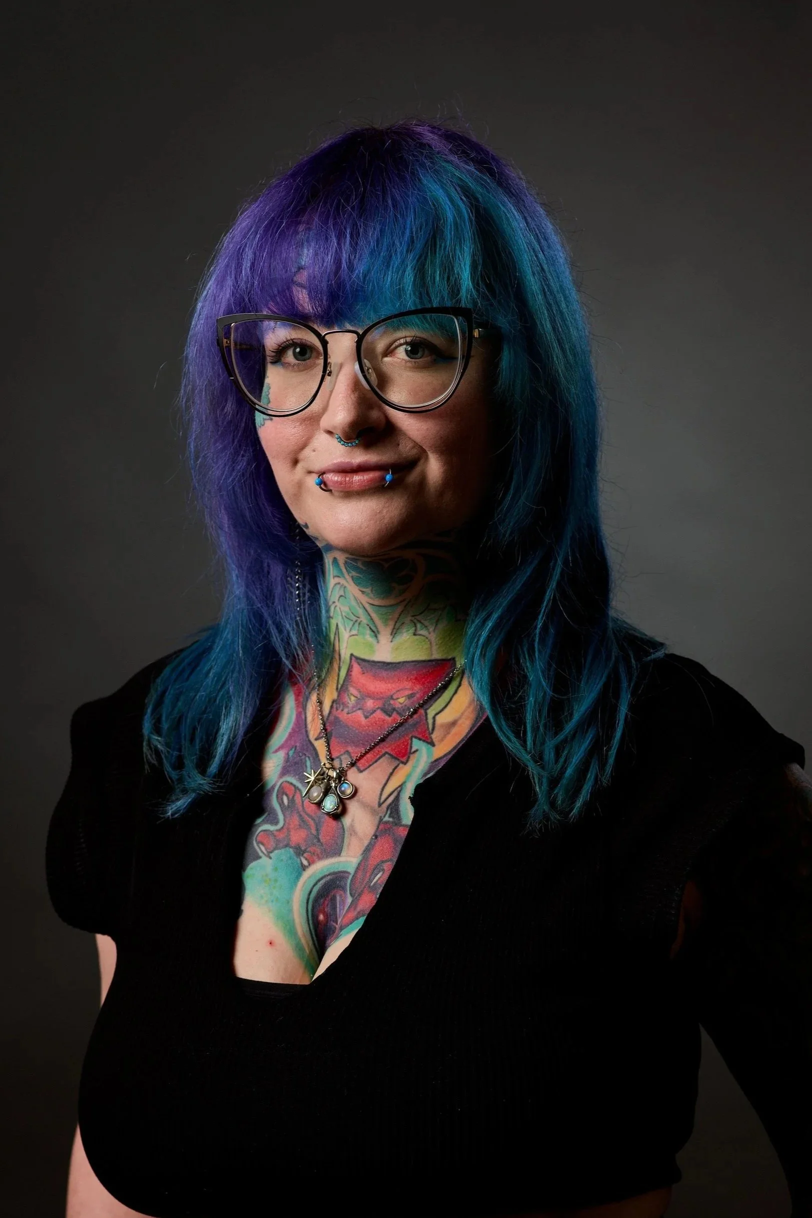 A woman with vibrant purple and blue hair, wearing glasses, tattoos on her chest and neck, and jewelry including a septum piercing and lip piercings, posing against a dark background.