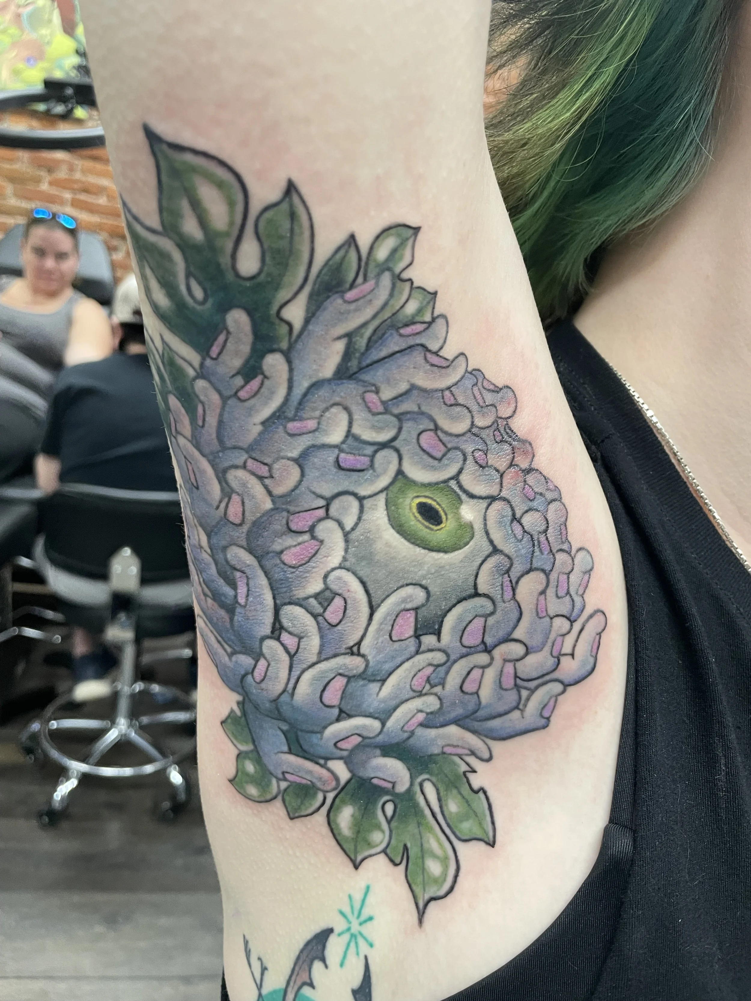 Close-up of a person's upper arm tattoo featuring a large, detailed green and purple floral design with leaves, and a small turquoise starburst beneath the design.