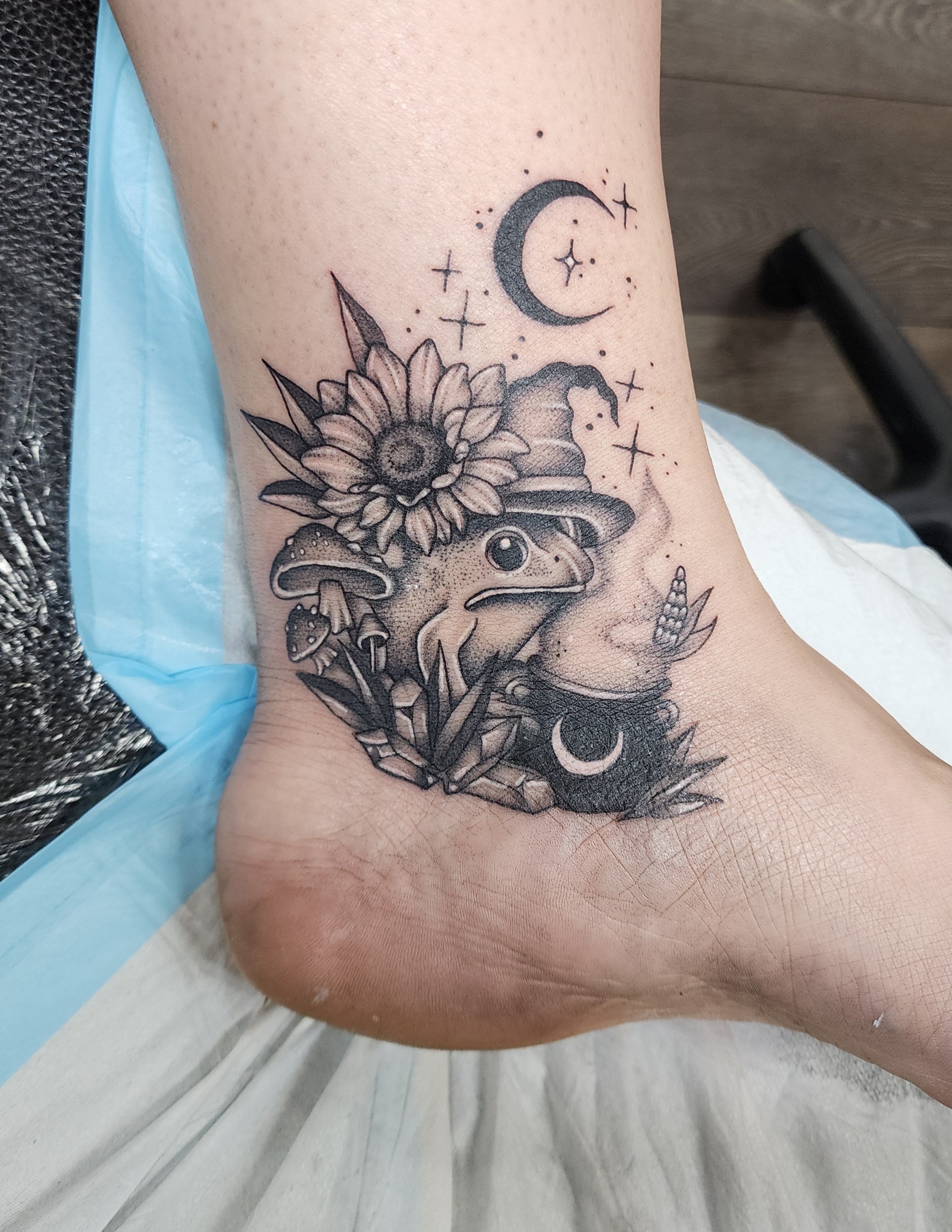 A tattoo on a person's arm featuring a sunflower, a crescent moon, and a small astronaut helmet with stars and celestial details.