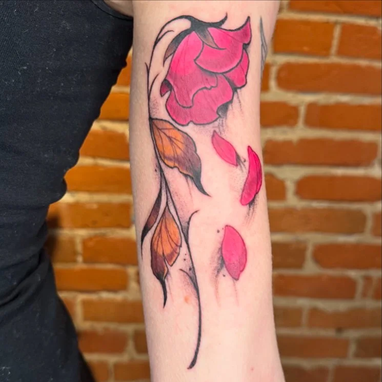 A colorful tattoo of a flower with pink petals, green and orange leaves, and a black stem on a person’s arm, with a brick wall background.