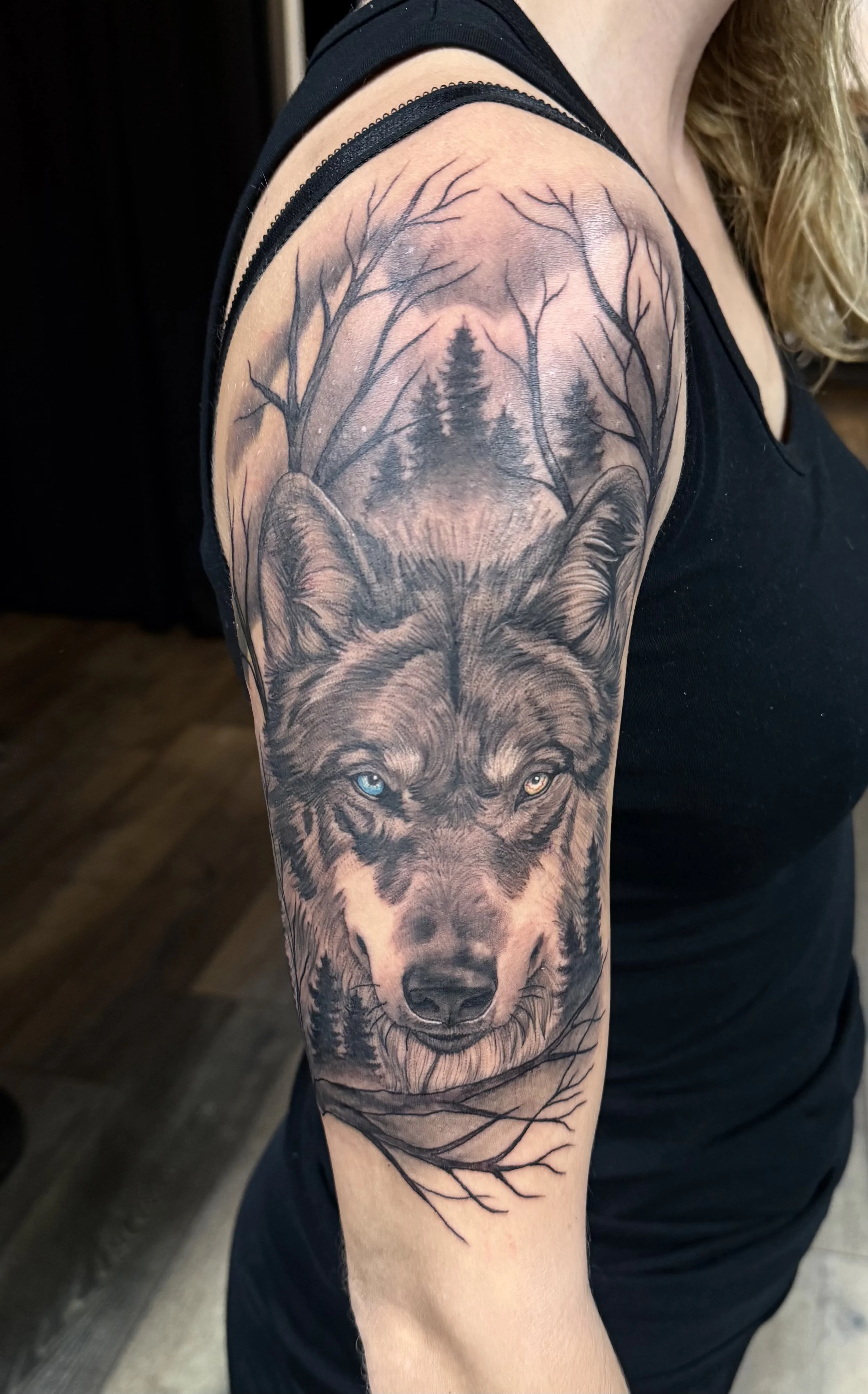Tattoo of a wolf's face with forest and bare trees in the background on a person's upper arm.