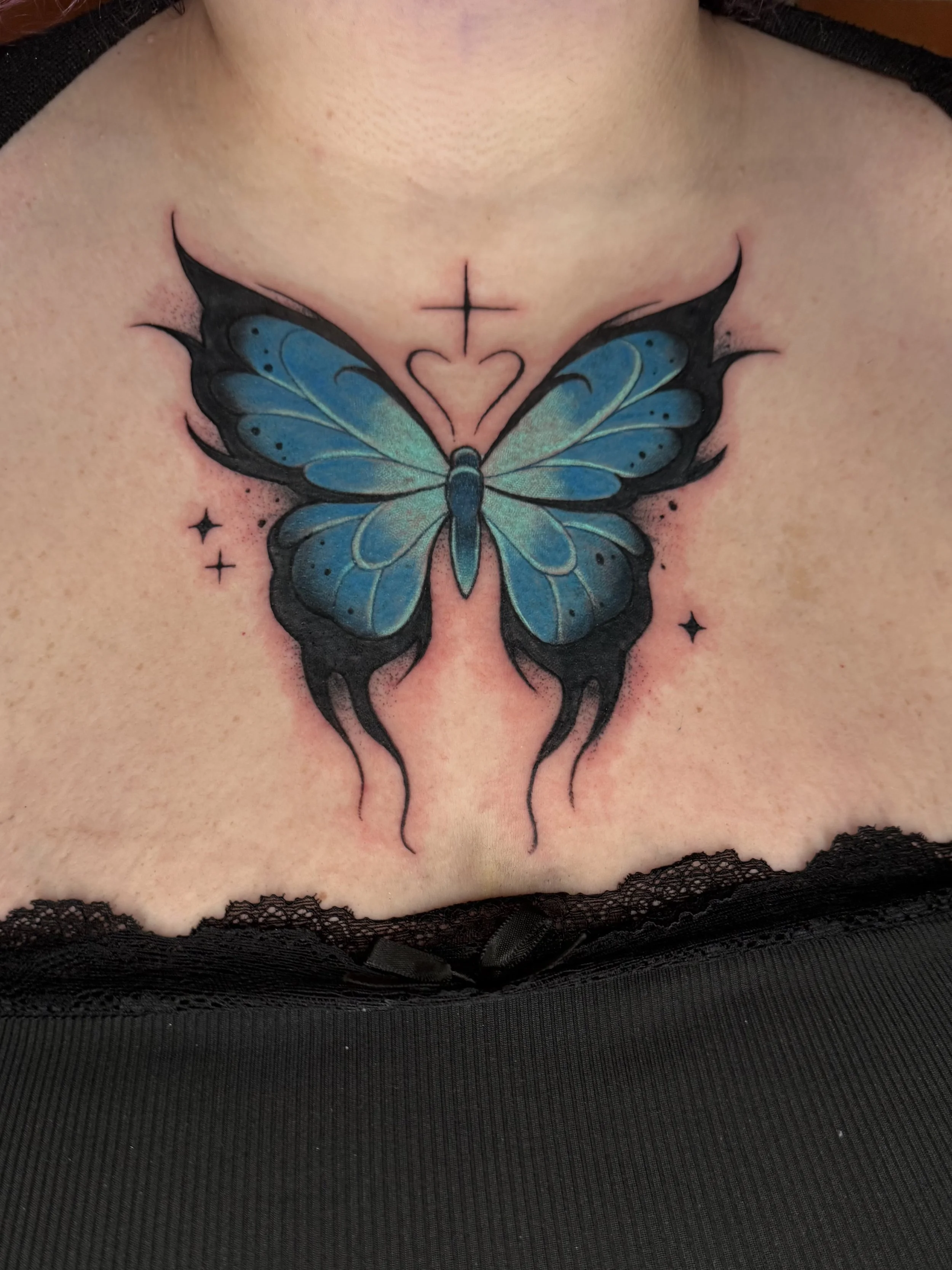A colorful tattoo of a blue butterfly on the upper chest, with a small cross and heart above it and three small stars around it, bordered by black lace fabric.