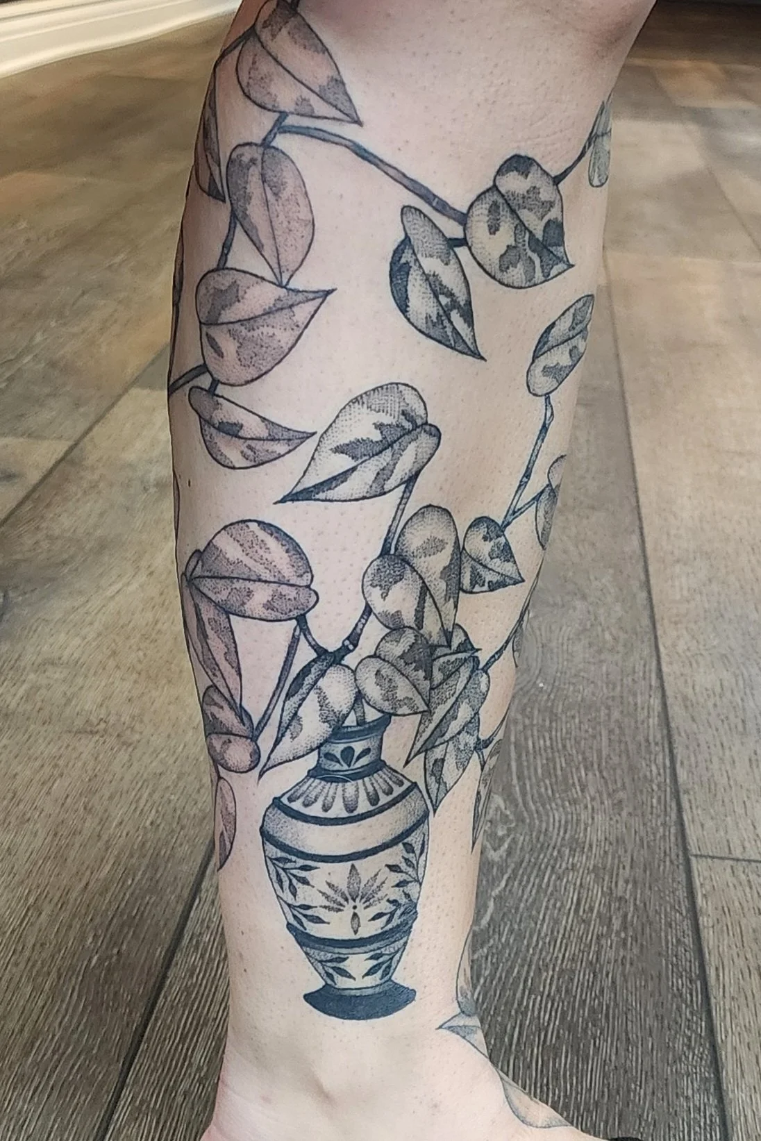 A person's leg with a black and gray tattoo of leaves on a vine in a vase.
