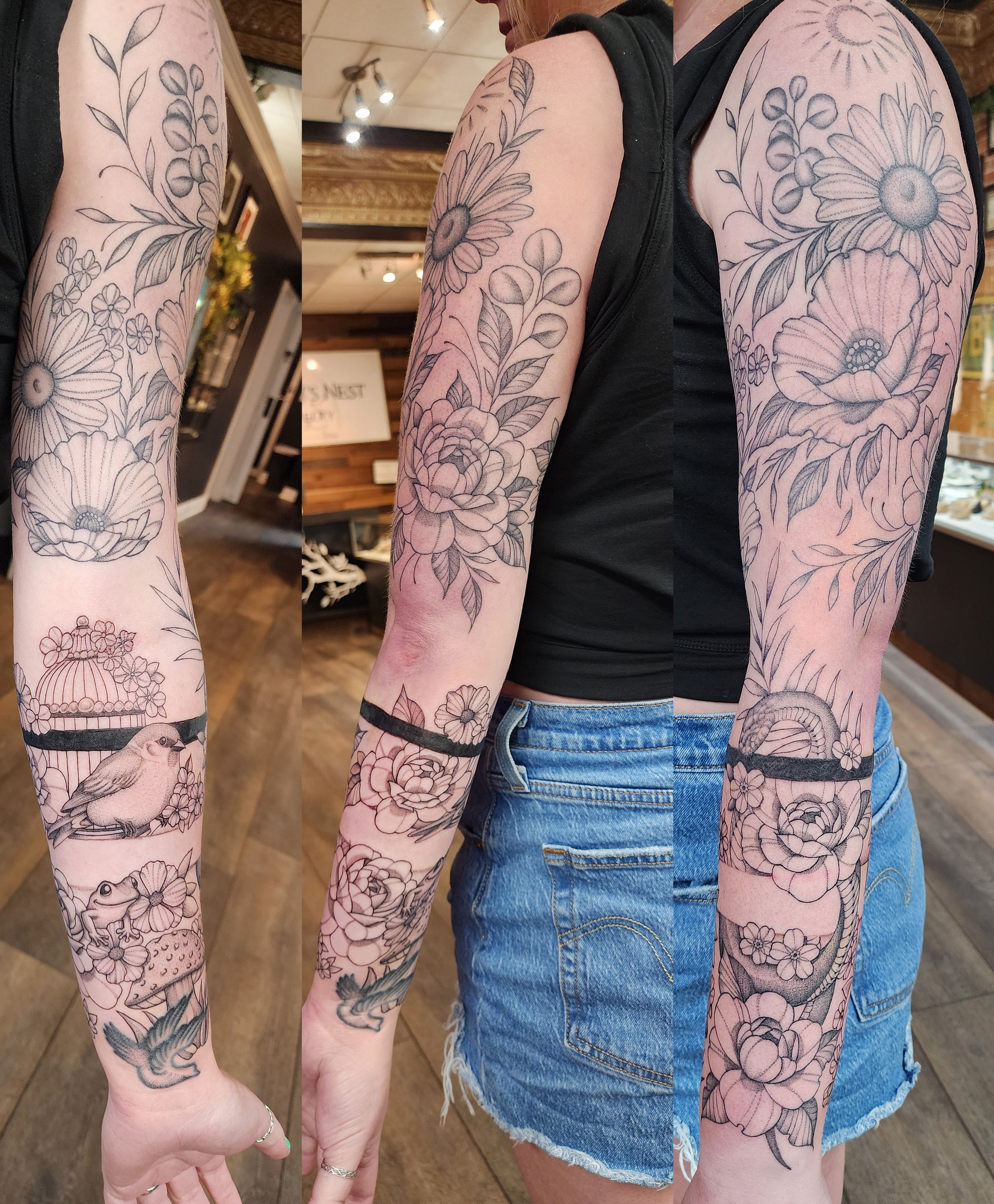 A person's arm with a detailed black and gray floral tattoo covering the entire arm, including sunflowers, roses, other flowers, leaves, and a bird perching on a branch, with intricate shading and line work.
