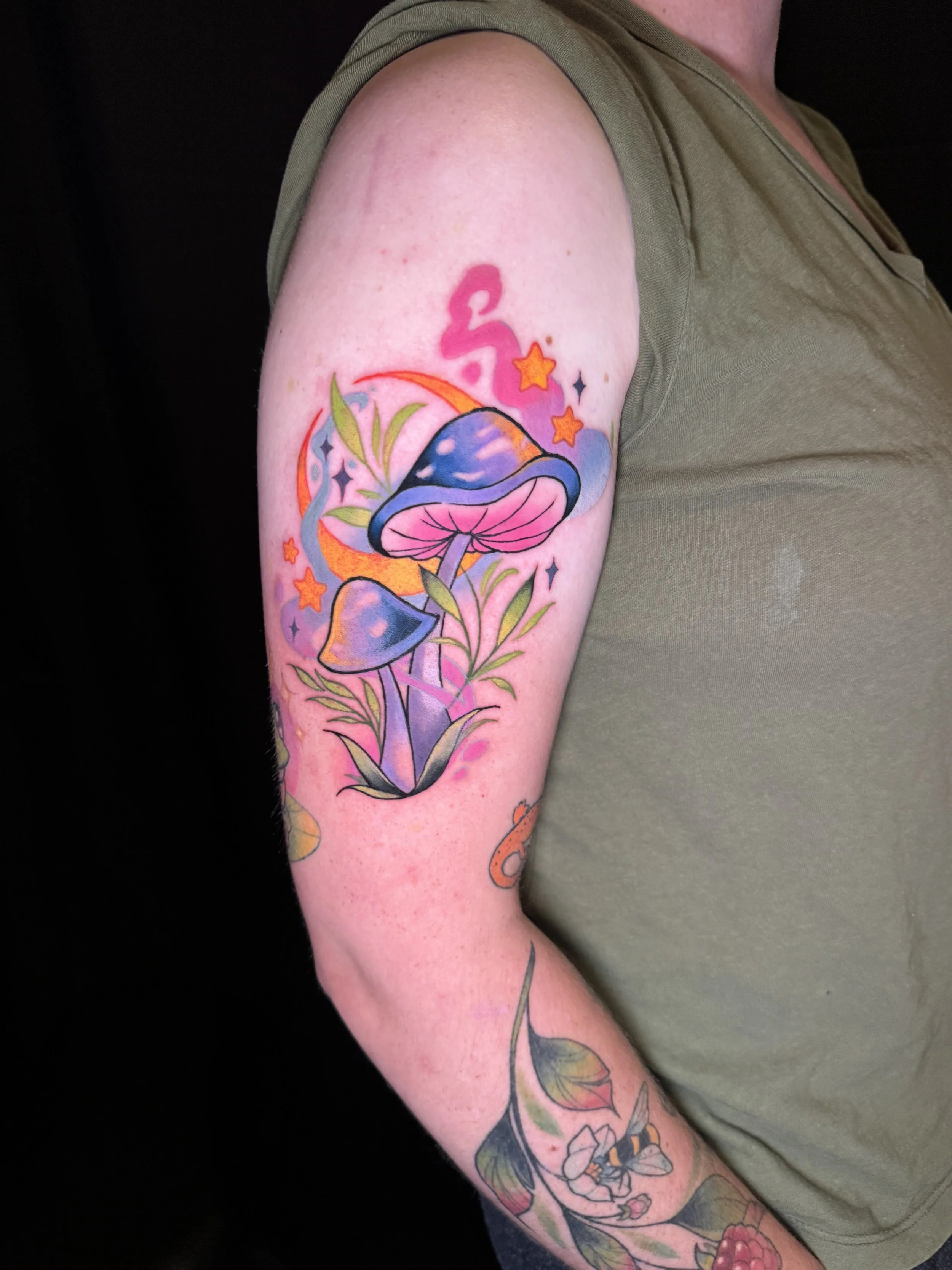 Colorful tattoo of a mushroom, stars, and botanical elements on person's upper arm, with part of the person's green shirt visible.