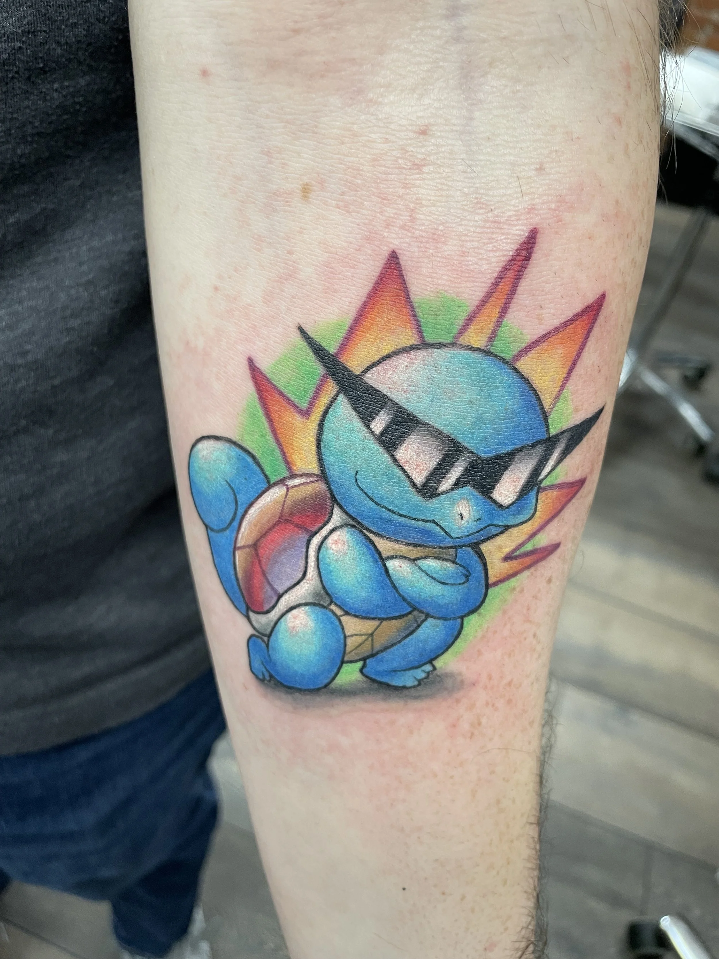 Colorful tattoo of a cute blue kitten with rainbow-colored sun rays and green background on a person's arm.