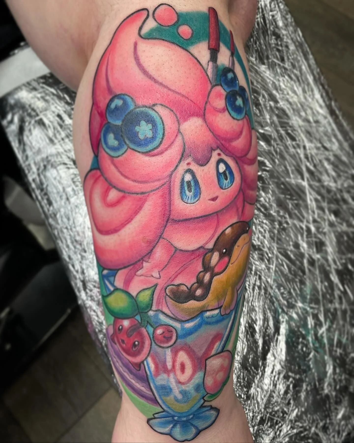 Colorful tattoo of Jigglypuff, a pink, round, Pokémon character with large blue eyes, surrounded by berries and leaves.