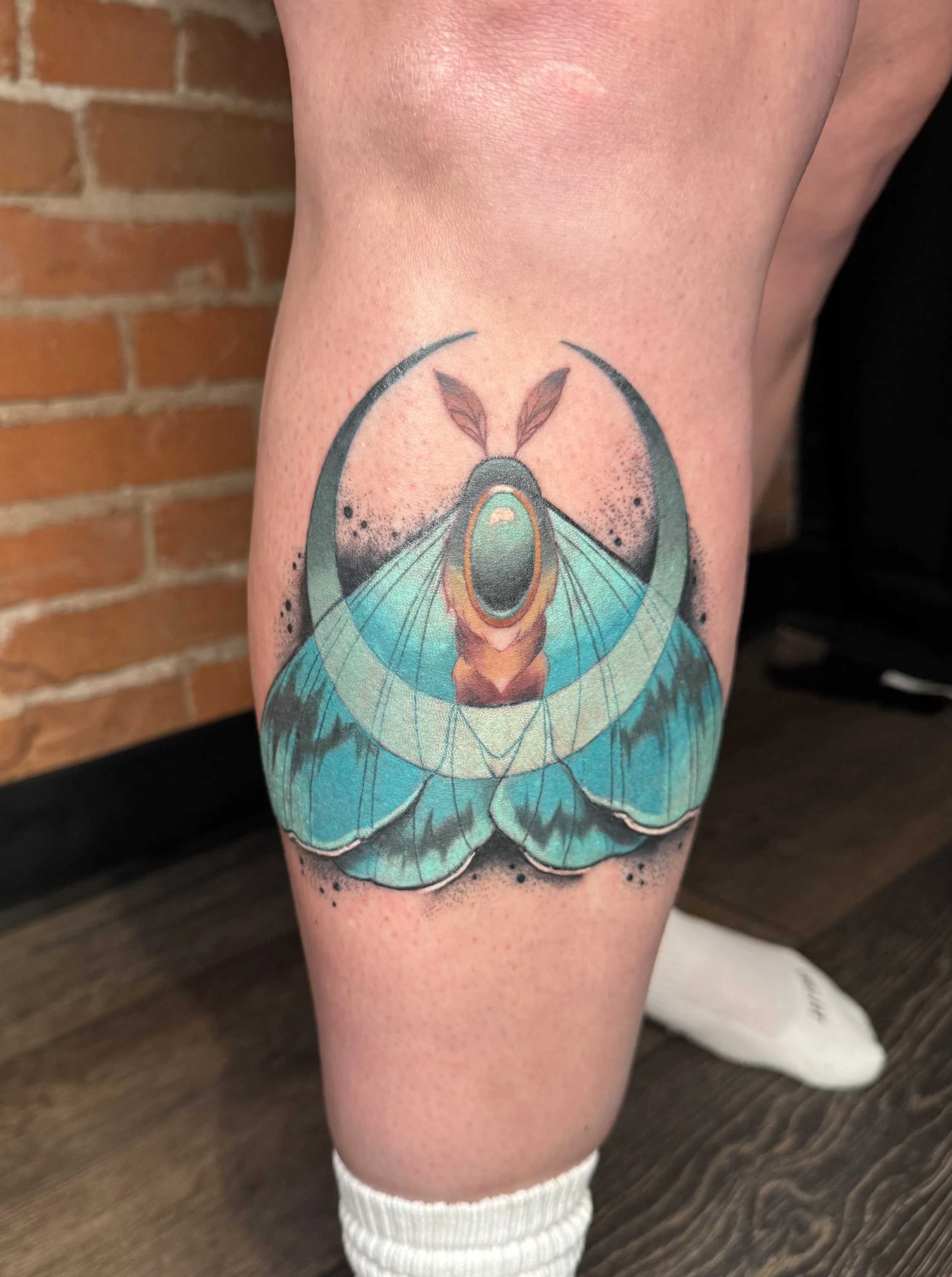Tattoo of a blue and orange moth on a person's thigh, with leaves and branches above its head, against a wooden background.