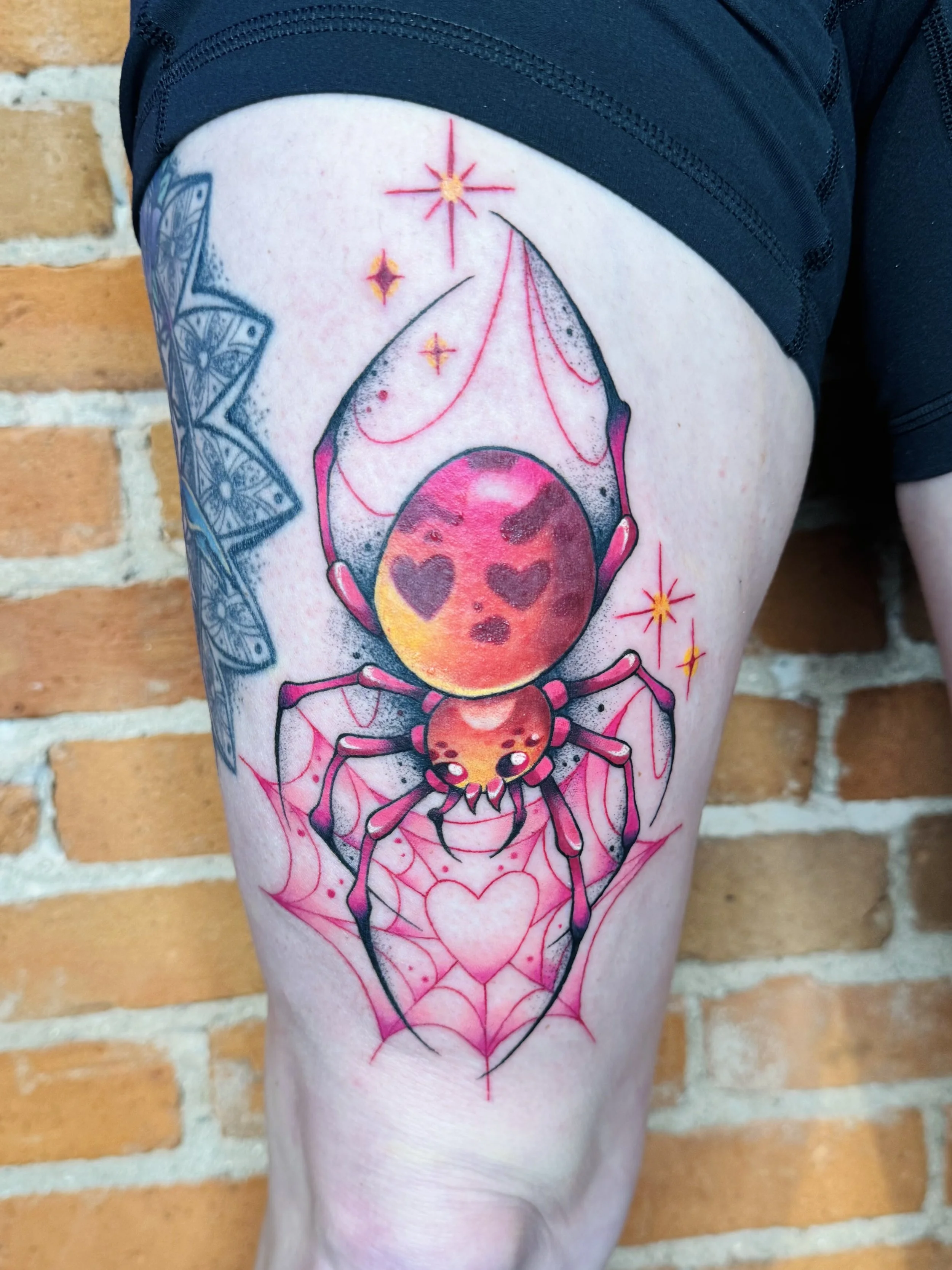 A colorful tattoo on a person's thigh featuring a spider holding a heart-shaped web with a heart inside it, with stars and sparkles surrounding the scene.