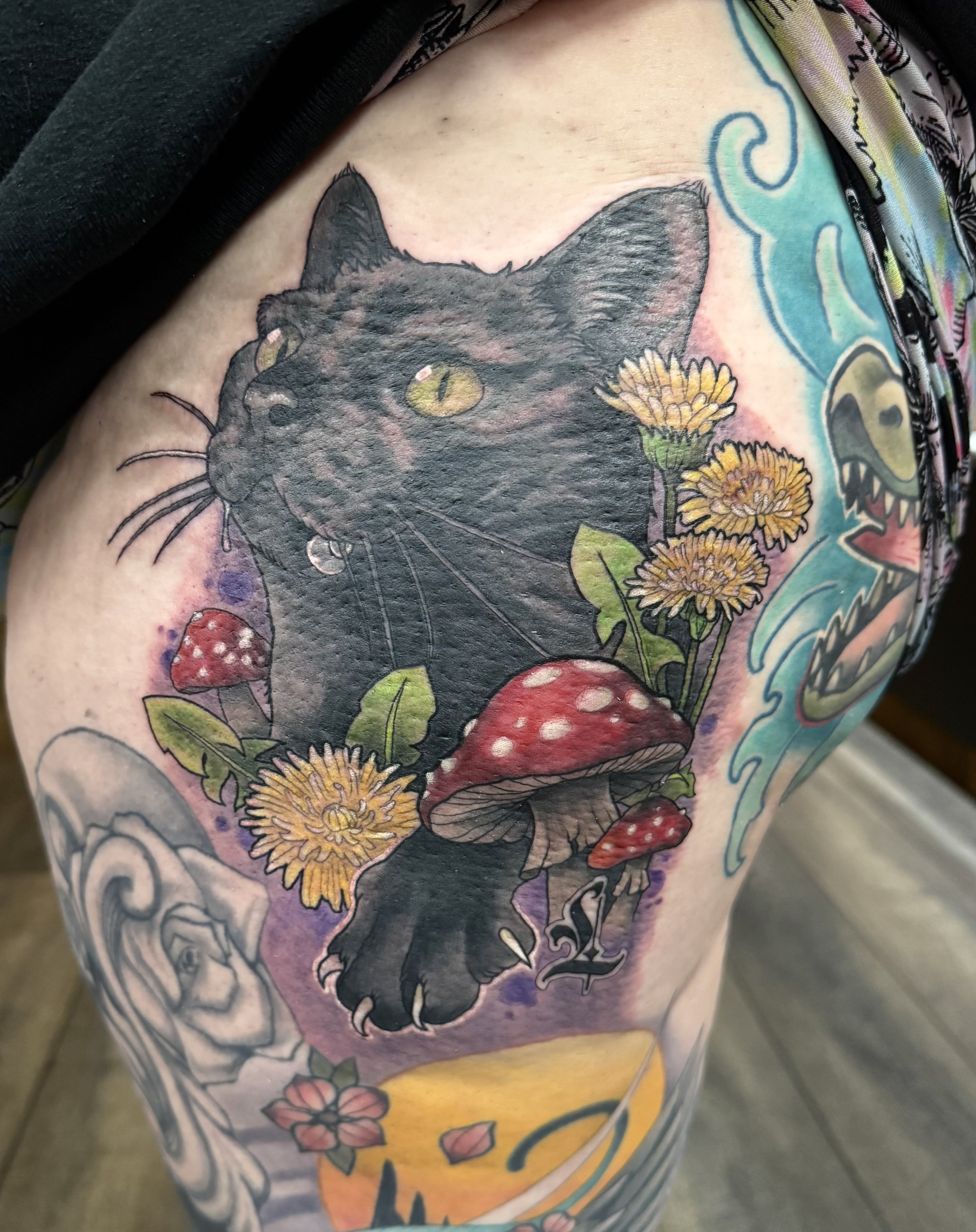 Colorful tattoo on a person's arm featuring a black cat's face with yellow eyes, surrounded by yellow flowers, mushrooms, and ocean wave designs with vibrant watercolor background.