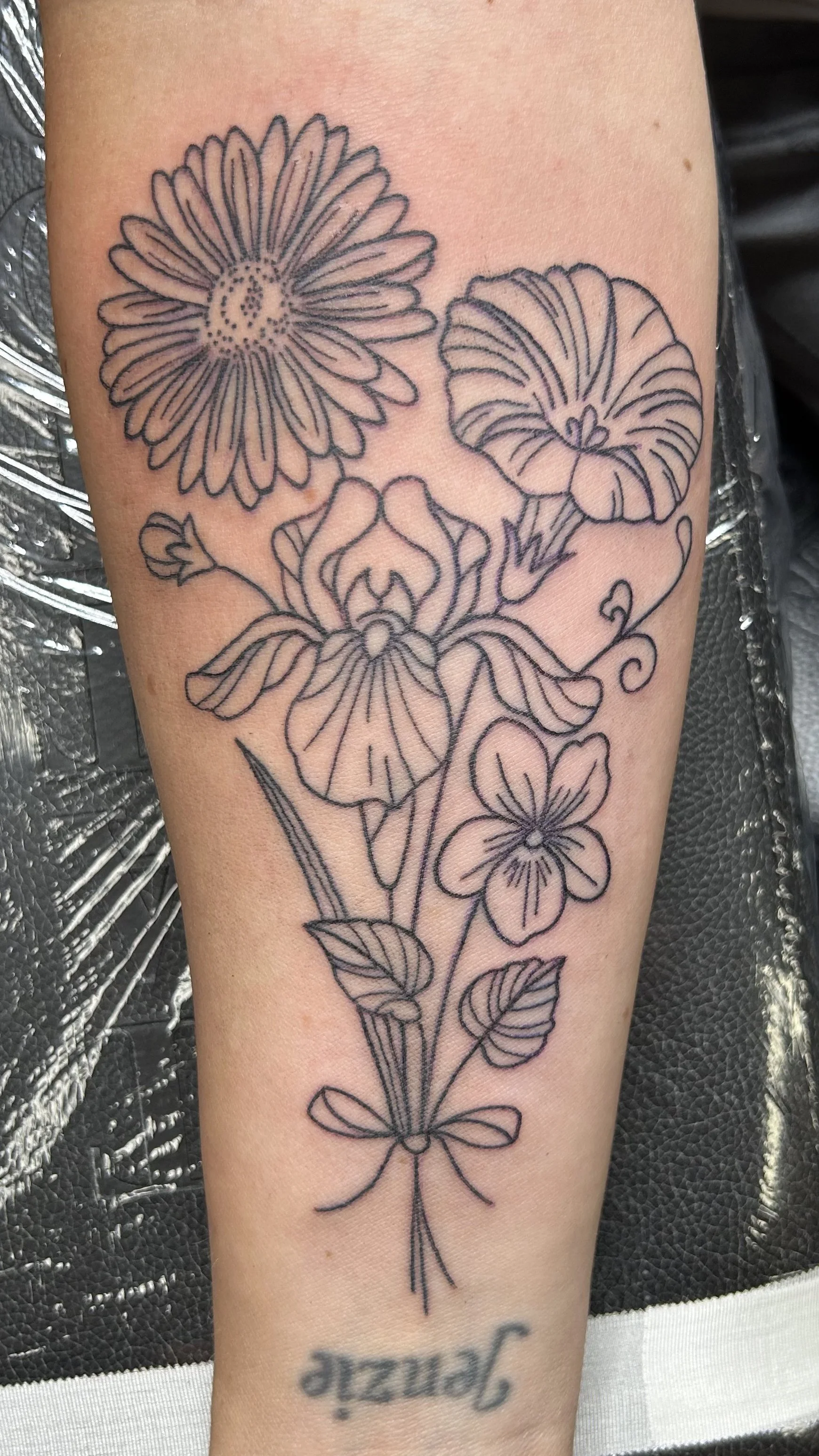 Line art tattoo of various flowers on skin, including daisies, pansies, and others, with leaves and stems.