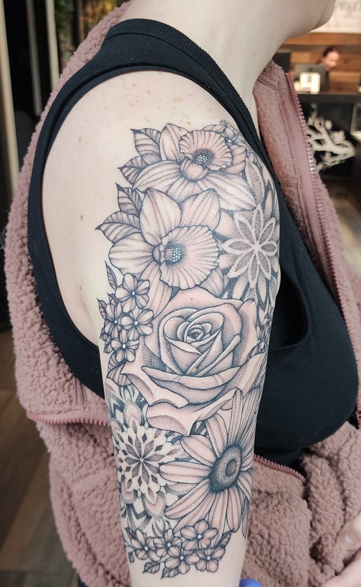 Black and gray floral tattoo on person's upper arm and shoulder, featuring roses, daisies, and other flowers in detailed line work.