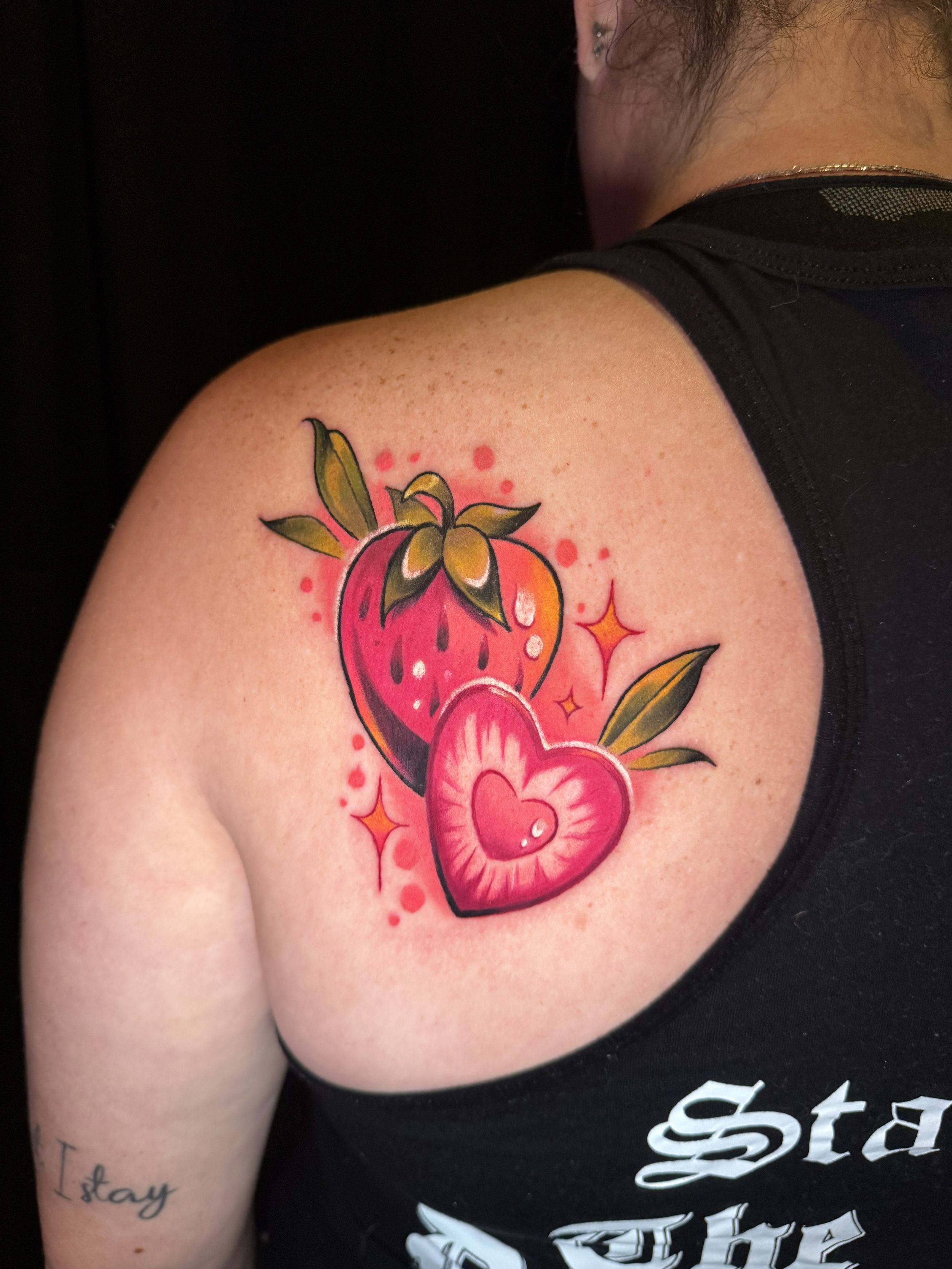 A person with a tattoo of a pink heart with a face, leaves, and stars on their upper arm.