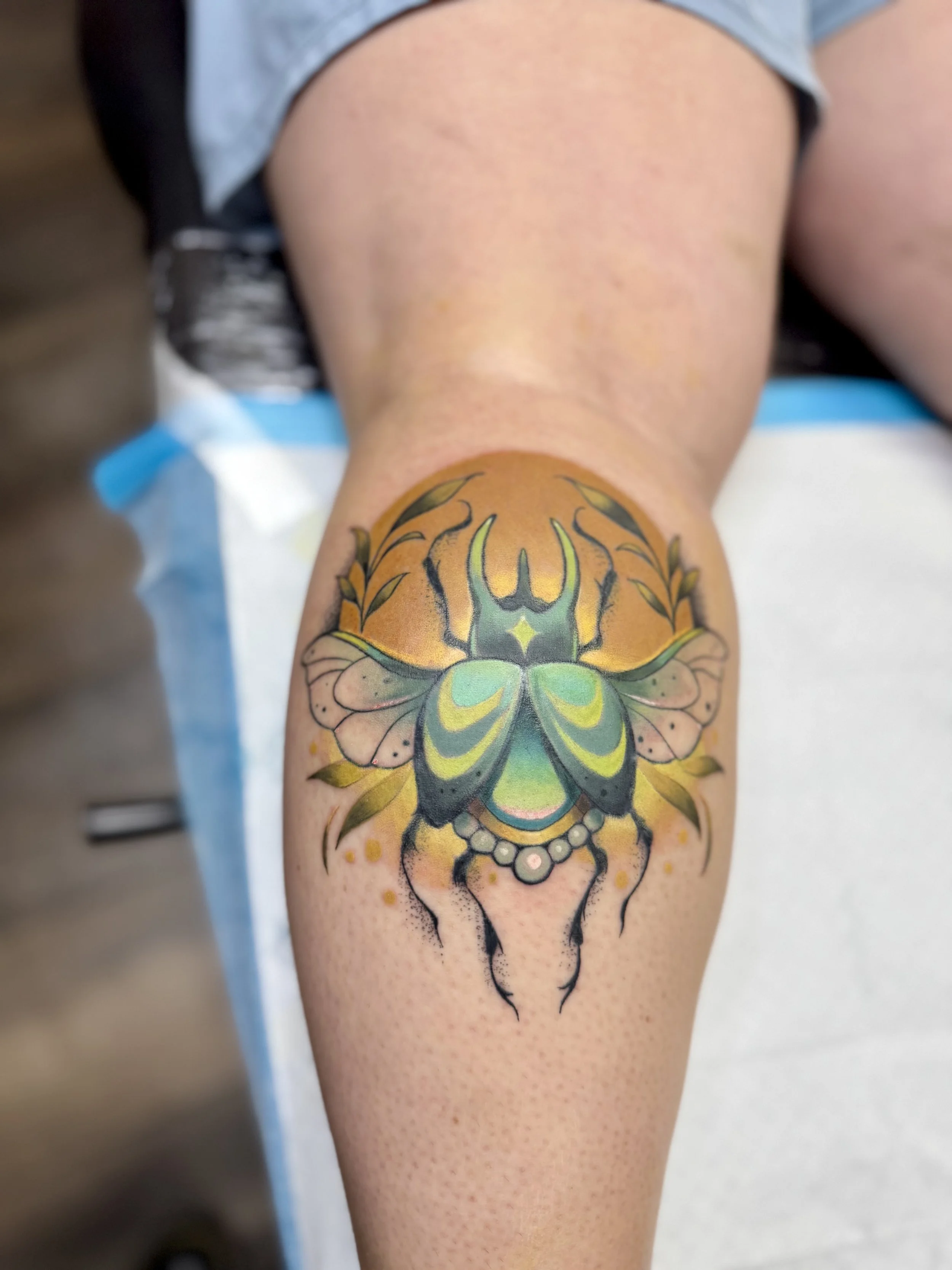 A person's thigh with a large, detailed tattoo of a colorful beetle with green, yellow, and black accents.