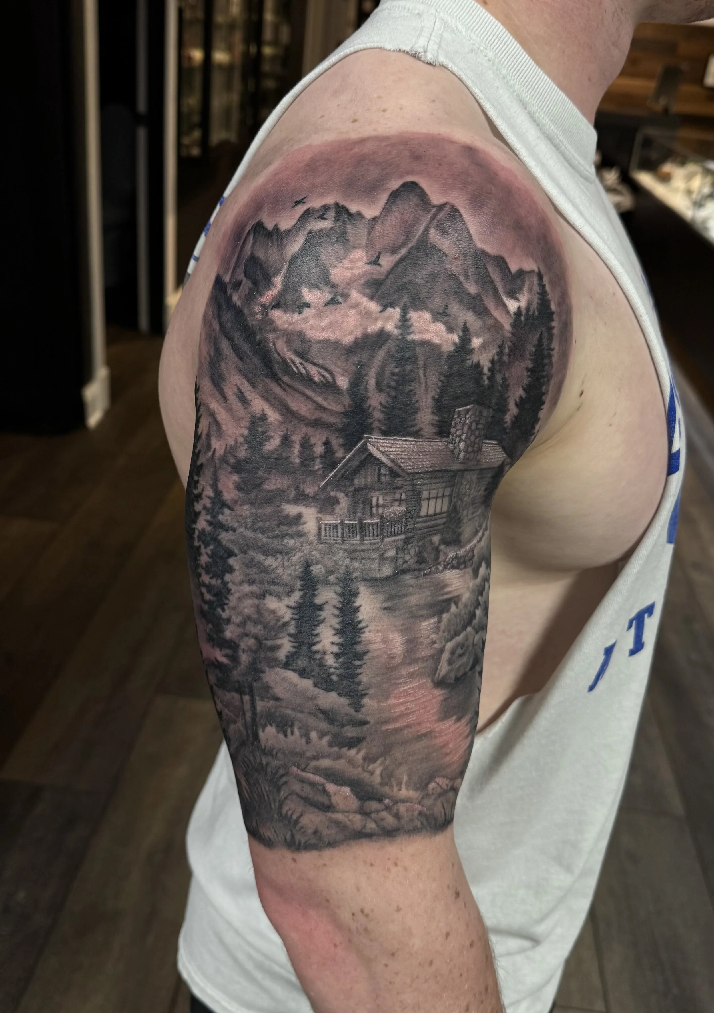 Black and gray landscape tattoo on a person's upper arm, depicting mountains, trees, a cabin with a chimney, and birds flying overhead.