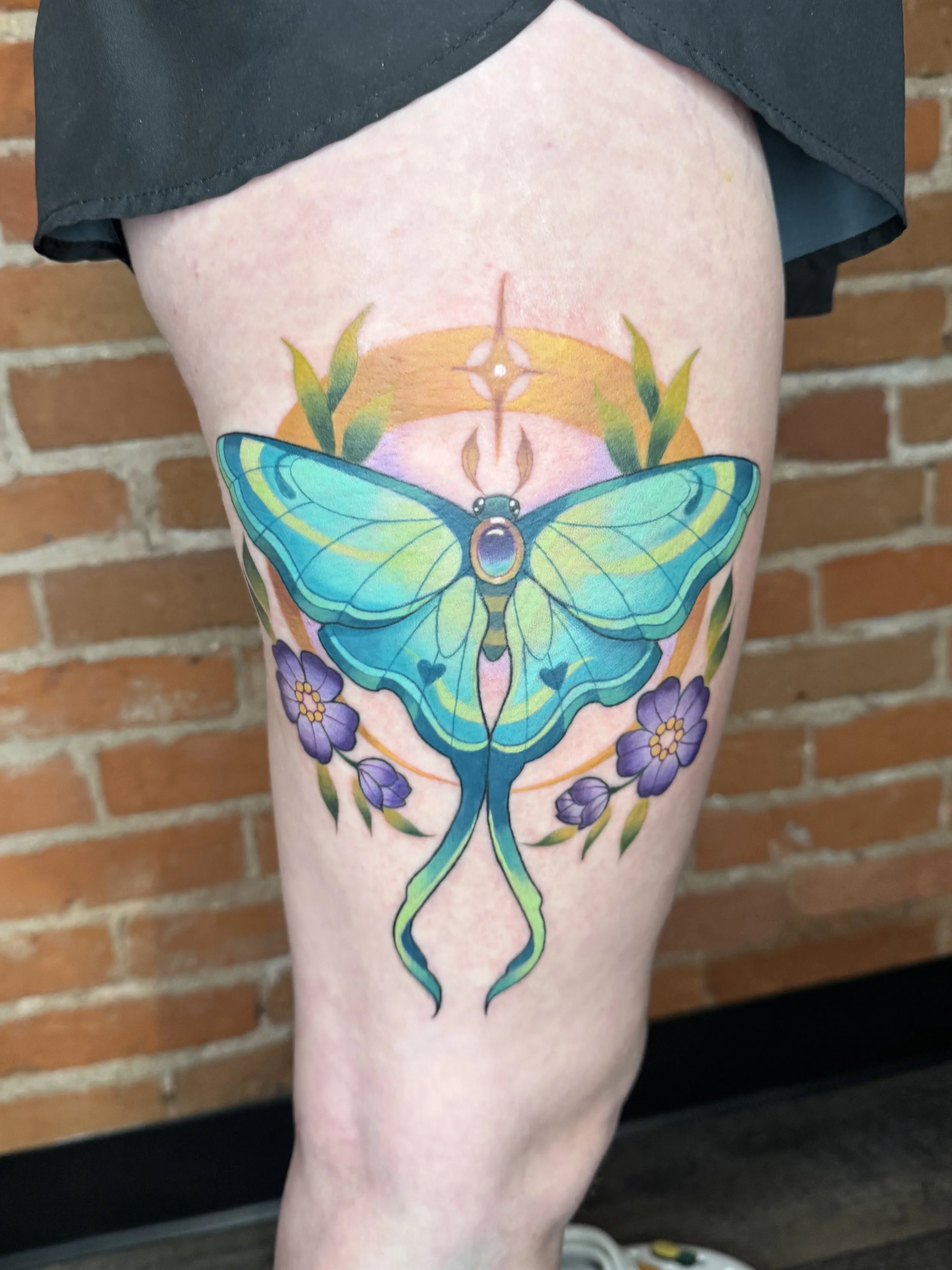 Colorful tattoo of a butterfly with purple flowers on the leg of a person standing in front of a brick wall.
