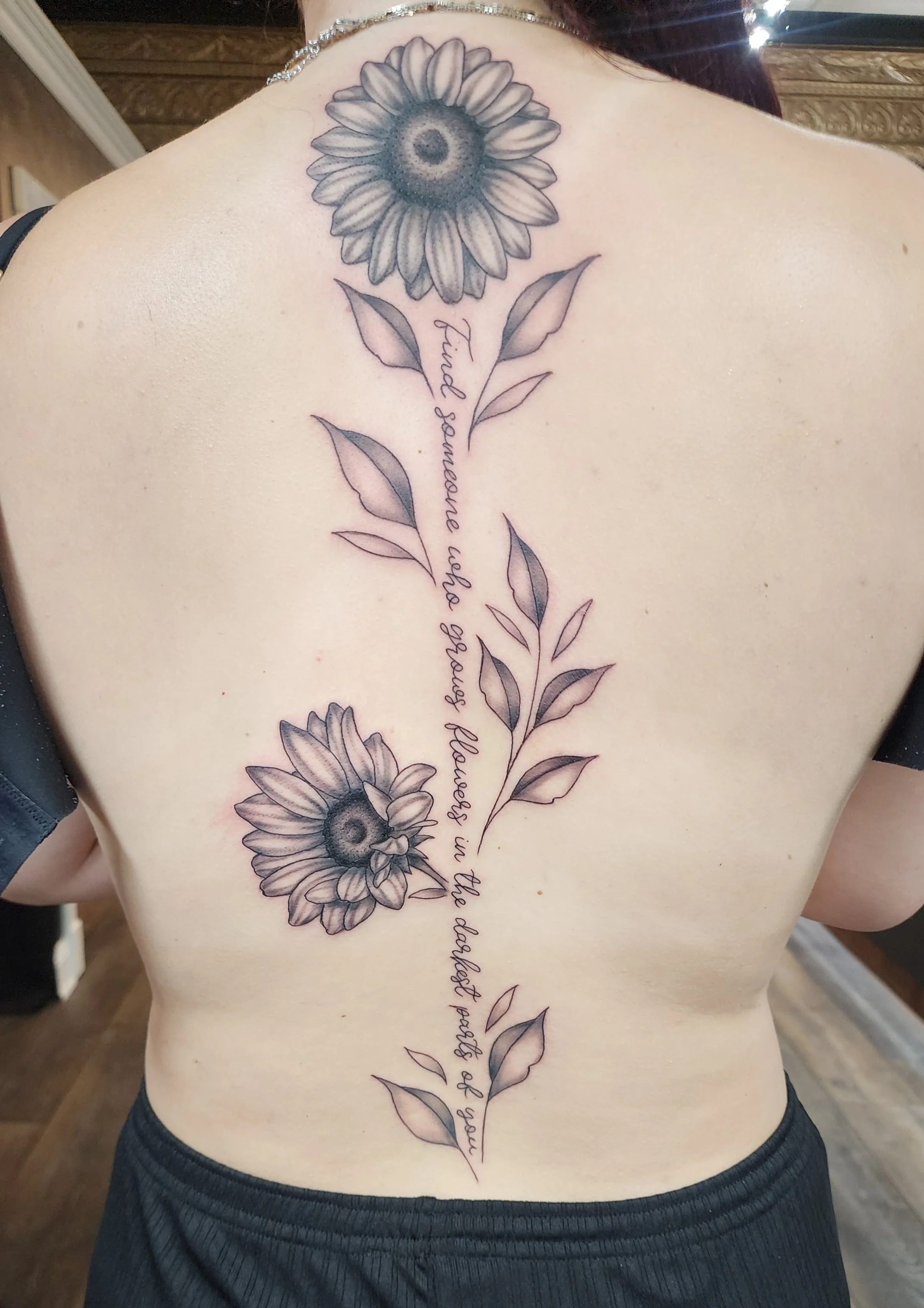 Tattoo of sunflowers with leaves and cursive text on a person's back.