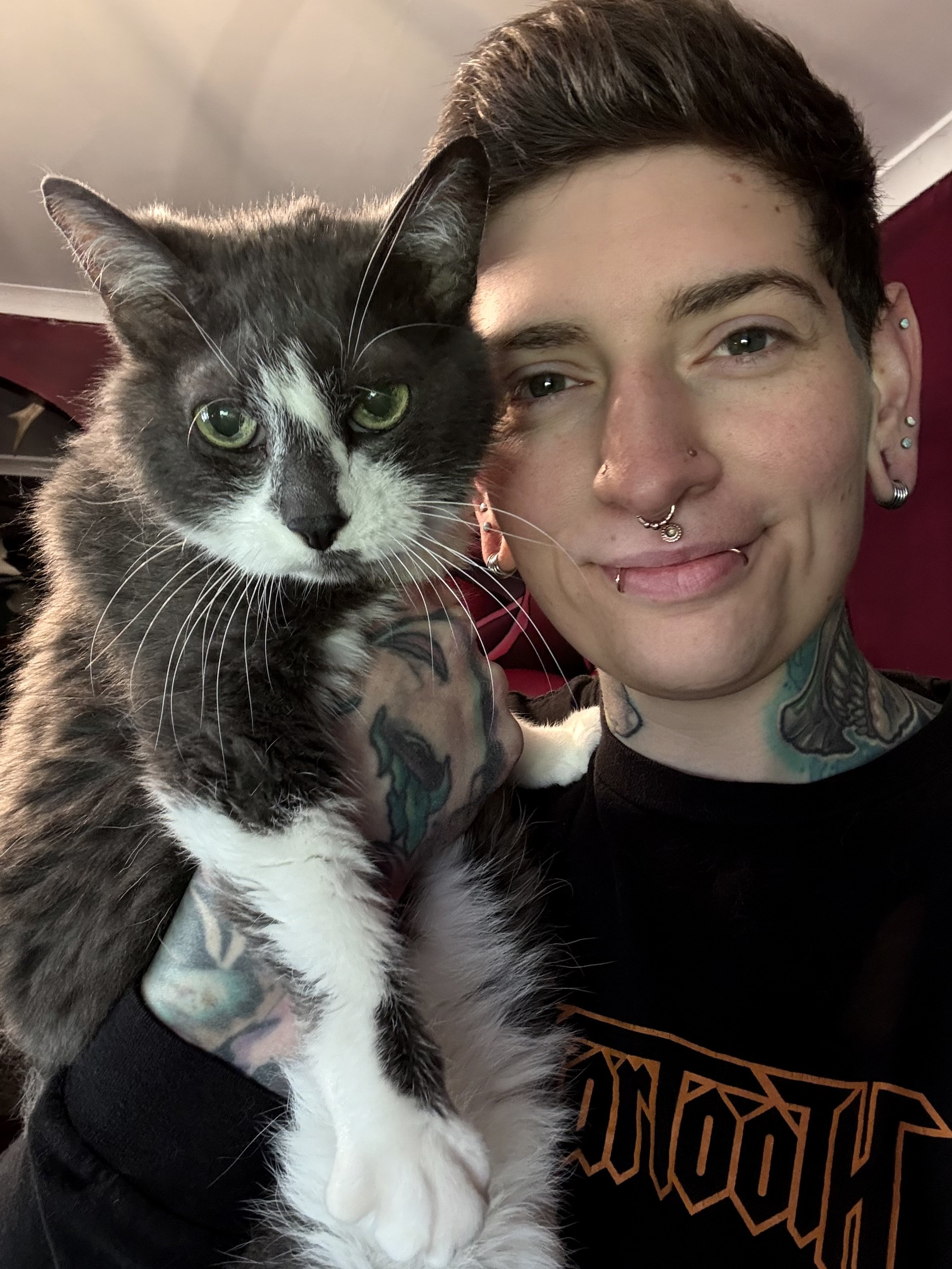 A person with short dark hair, multiple facial piercings, tattoos, and a smile, sitting close with a gray and white cat with green eyes, in a room with a red background.