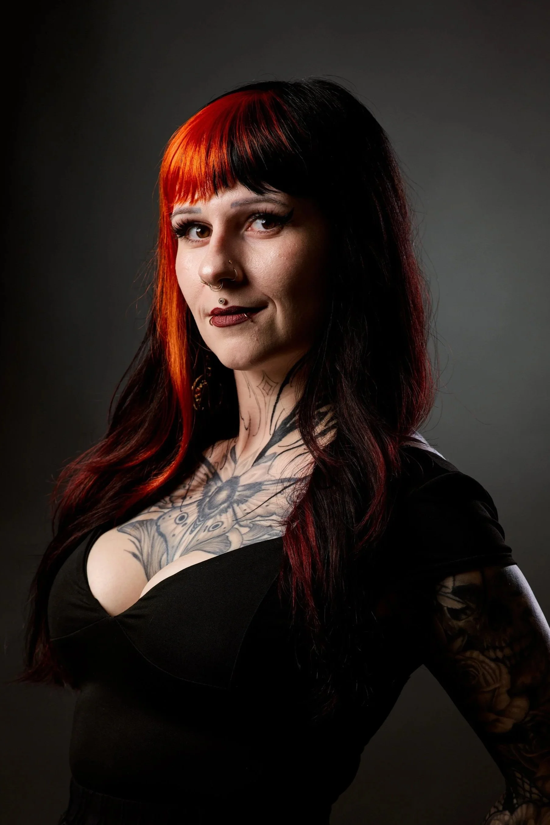 A woman with black and red hair, tattoos on her chest and arms, wearing a black top, looking at the camera, with a dark gray background.