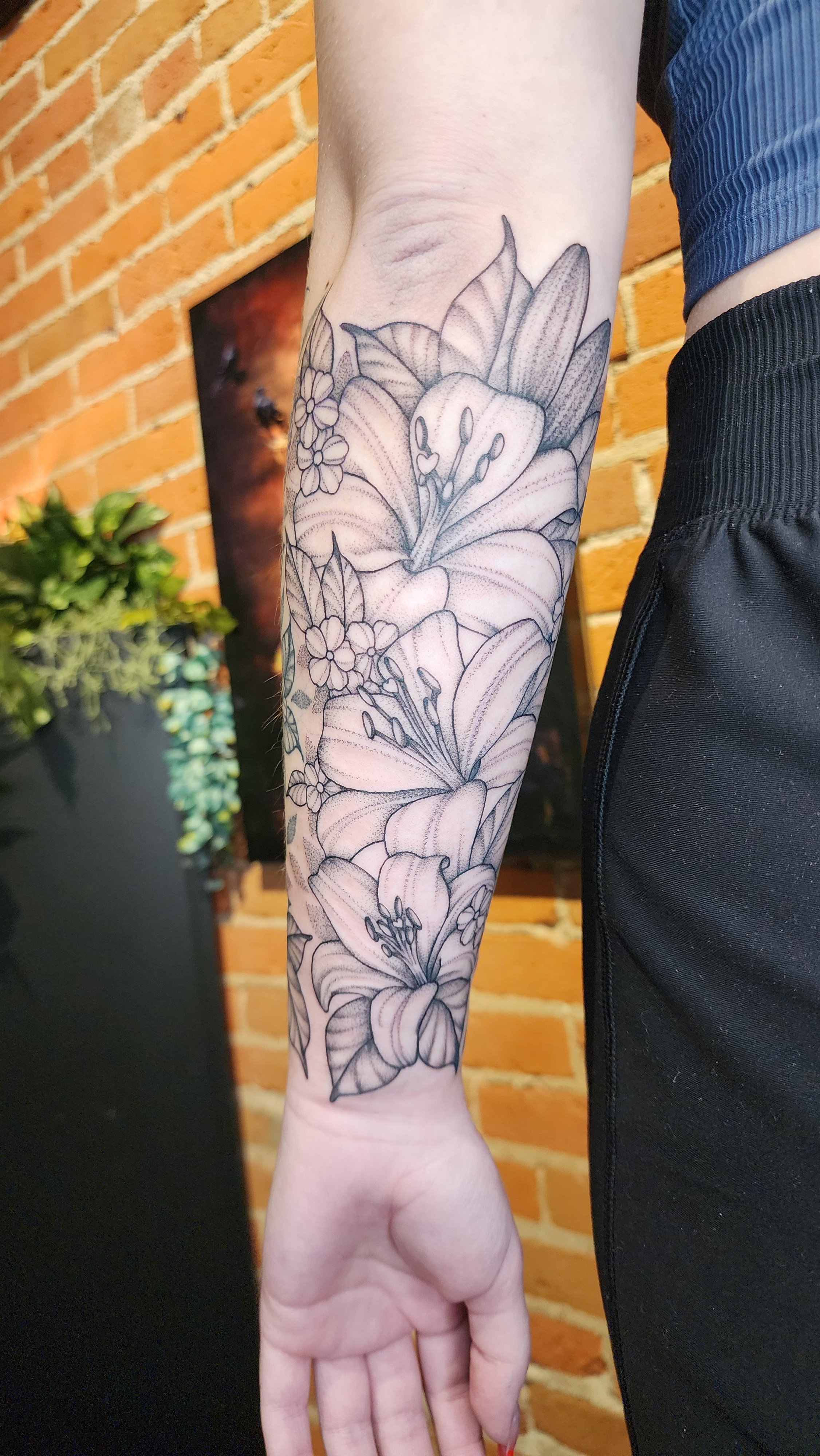 Black and gray floral tattoo on person's forearm with large lilies and smaller flowers, against a brick wall background.