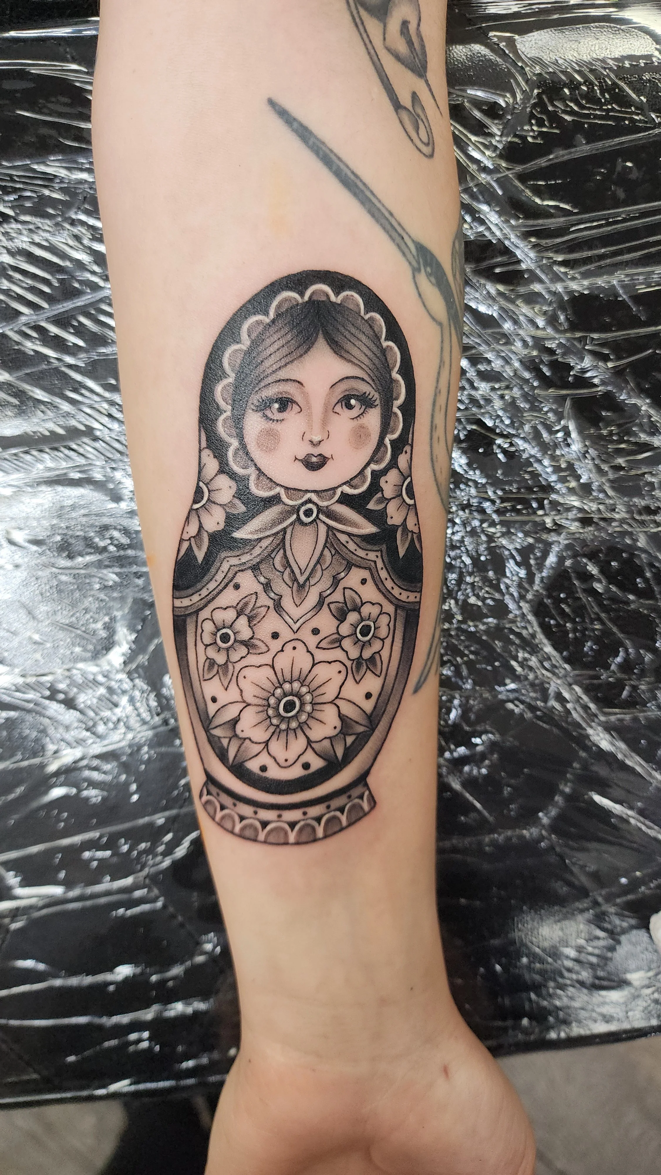 Tattoo of a stylized female face with large eyes, rosy cheeks, and black hair, surrounded by floral patterns in black ink on the person's arm.