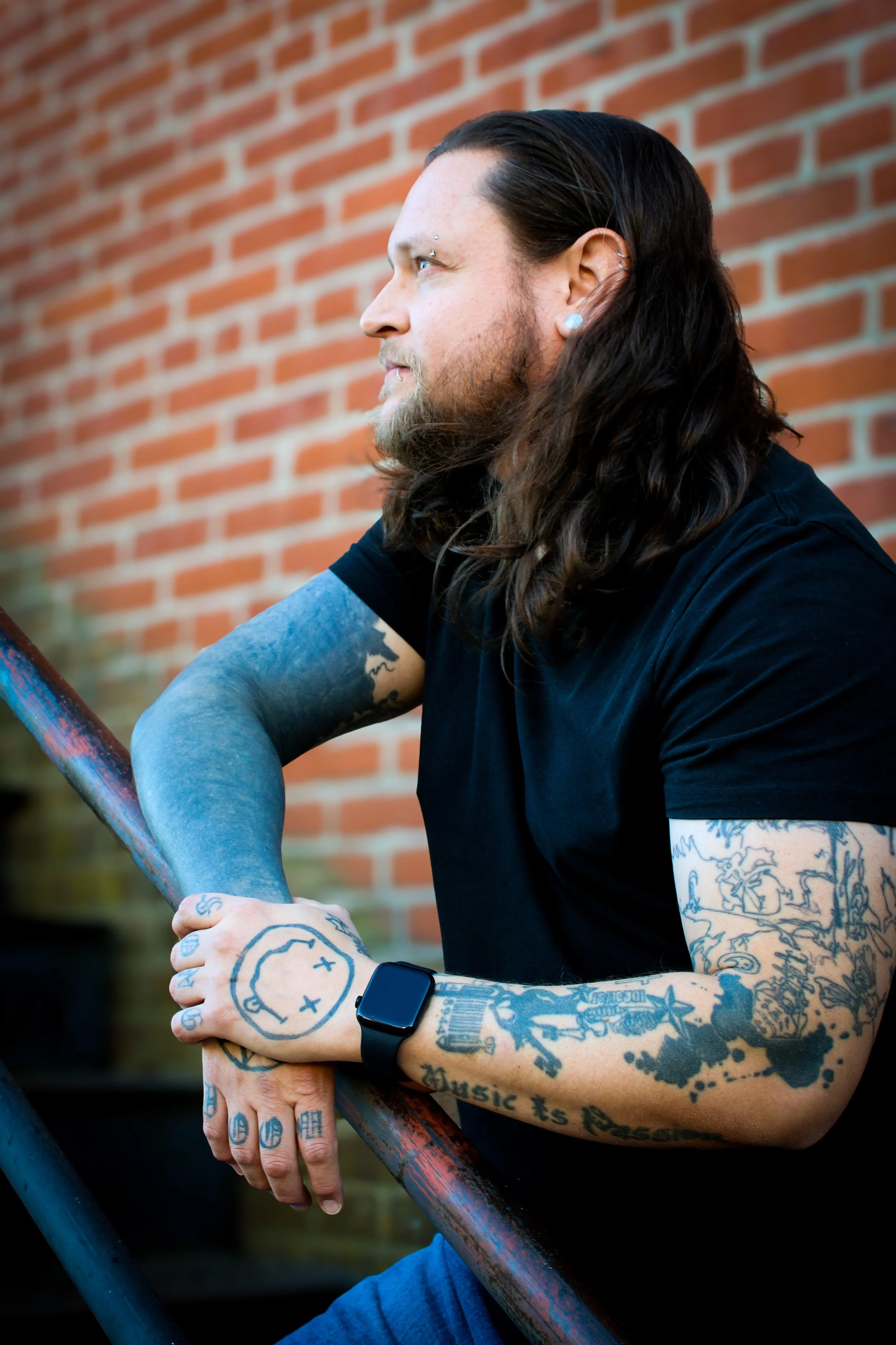 A man with long dark hair and tattoos on his arms and hands, wearing a black T-shirt and a smartwatch, sitting outdoors against a brick wall, resting his arms on a metal railing.