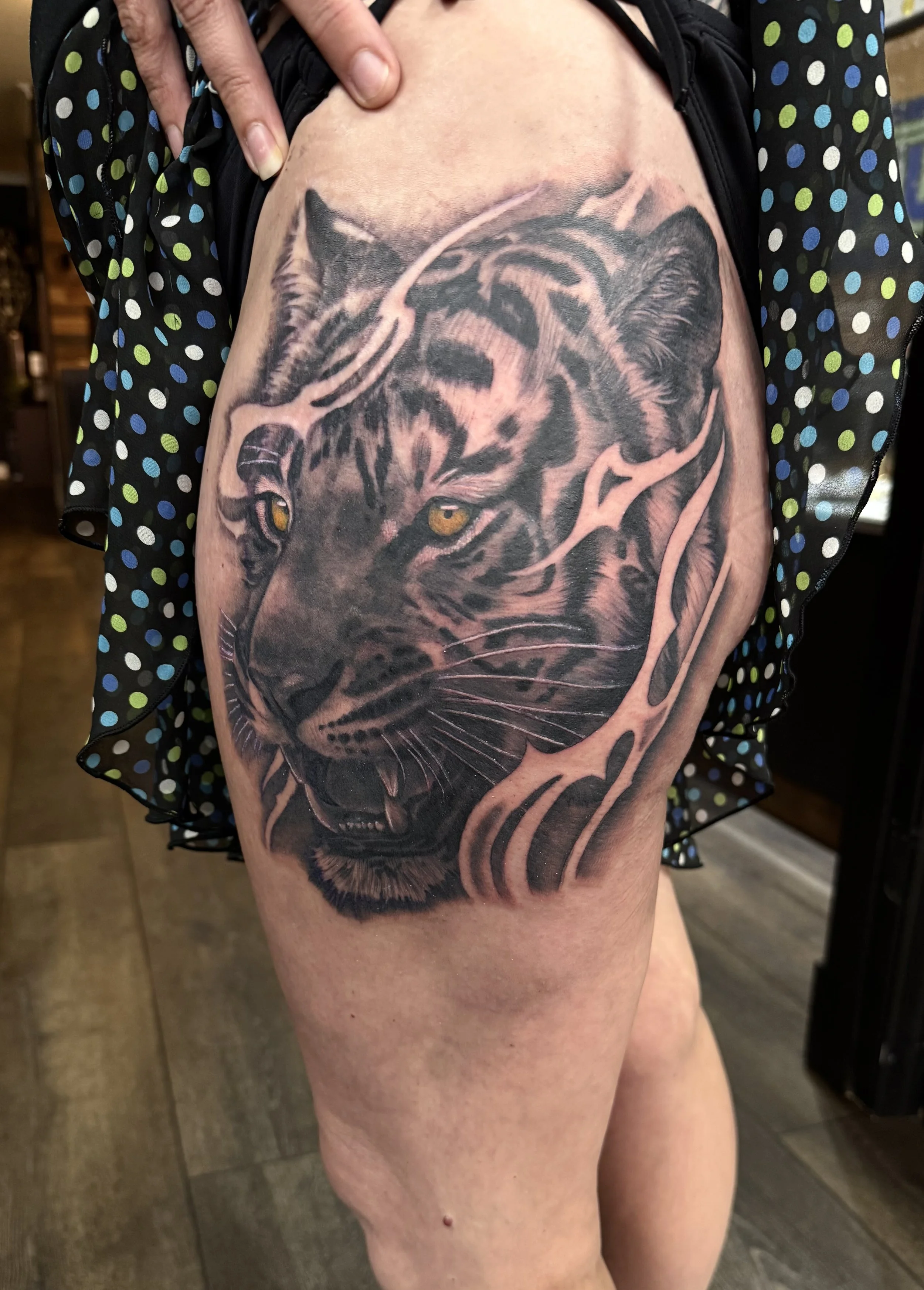 Tattoo of a black and gray tiger face with piercing yellow eyes on a person's upper thigh.
