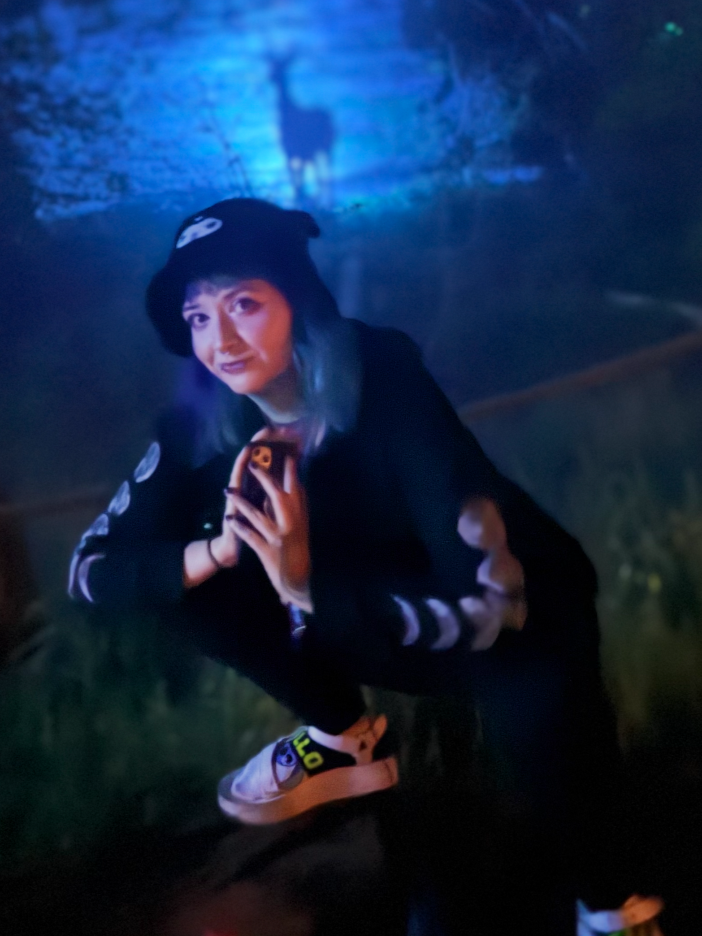 A young woman crouching in a dimly lit outdoor setting at night, wearing a black hoodie with white graphics, a black beanie with a skull design, holding her phone, and smiling at the camera.