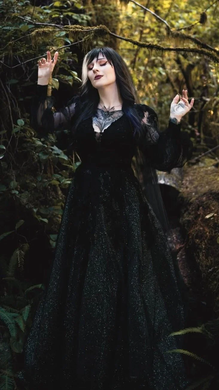 A woman in a black, glittery gown standing in a forest with her eyes closed, arms raised, surrounded by green foliage and moss-covered trees.