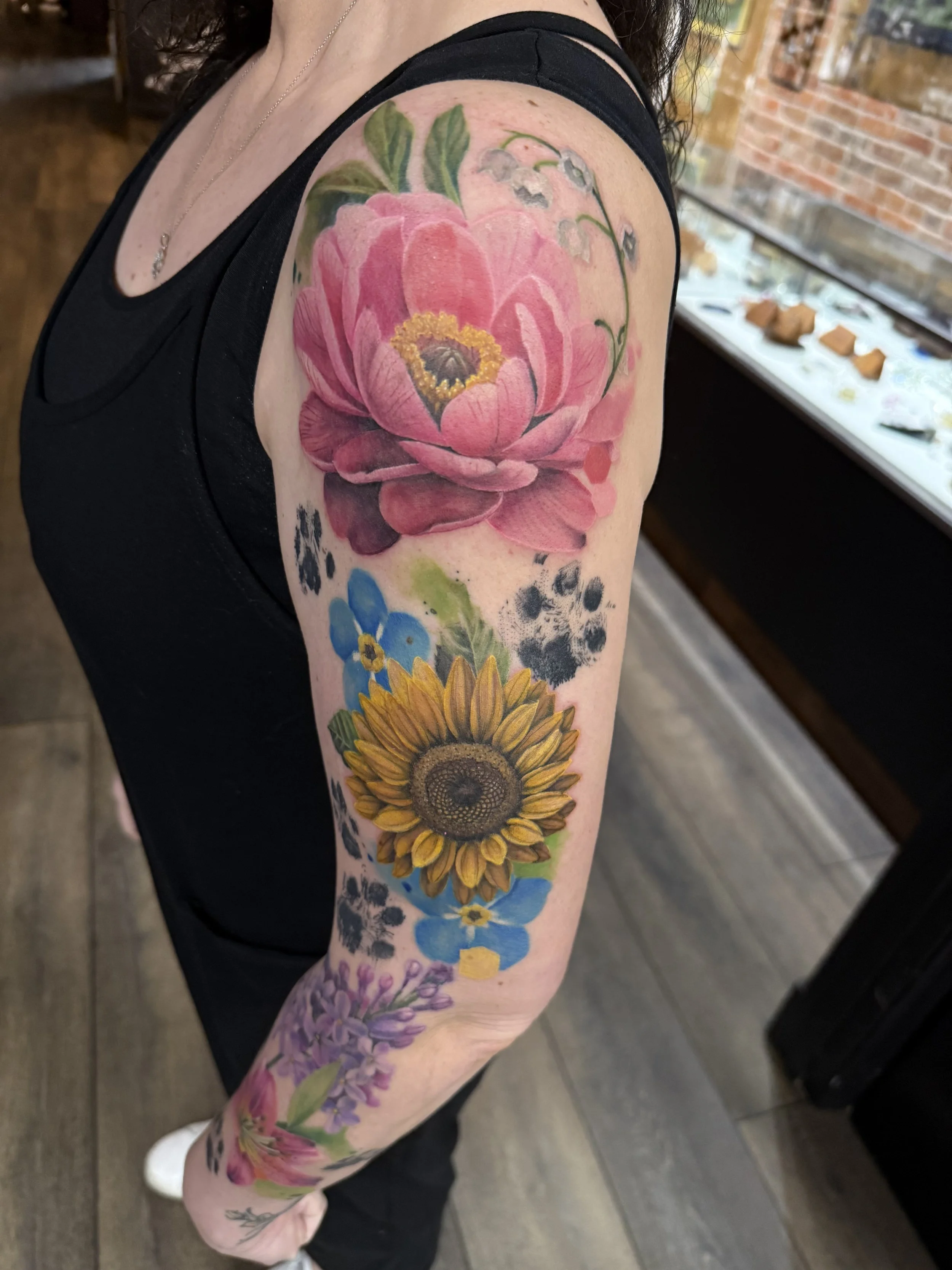 A person's arm with a colorful floral tattoo featuring a pink peony, sunflower, and other flowers and leaves.