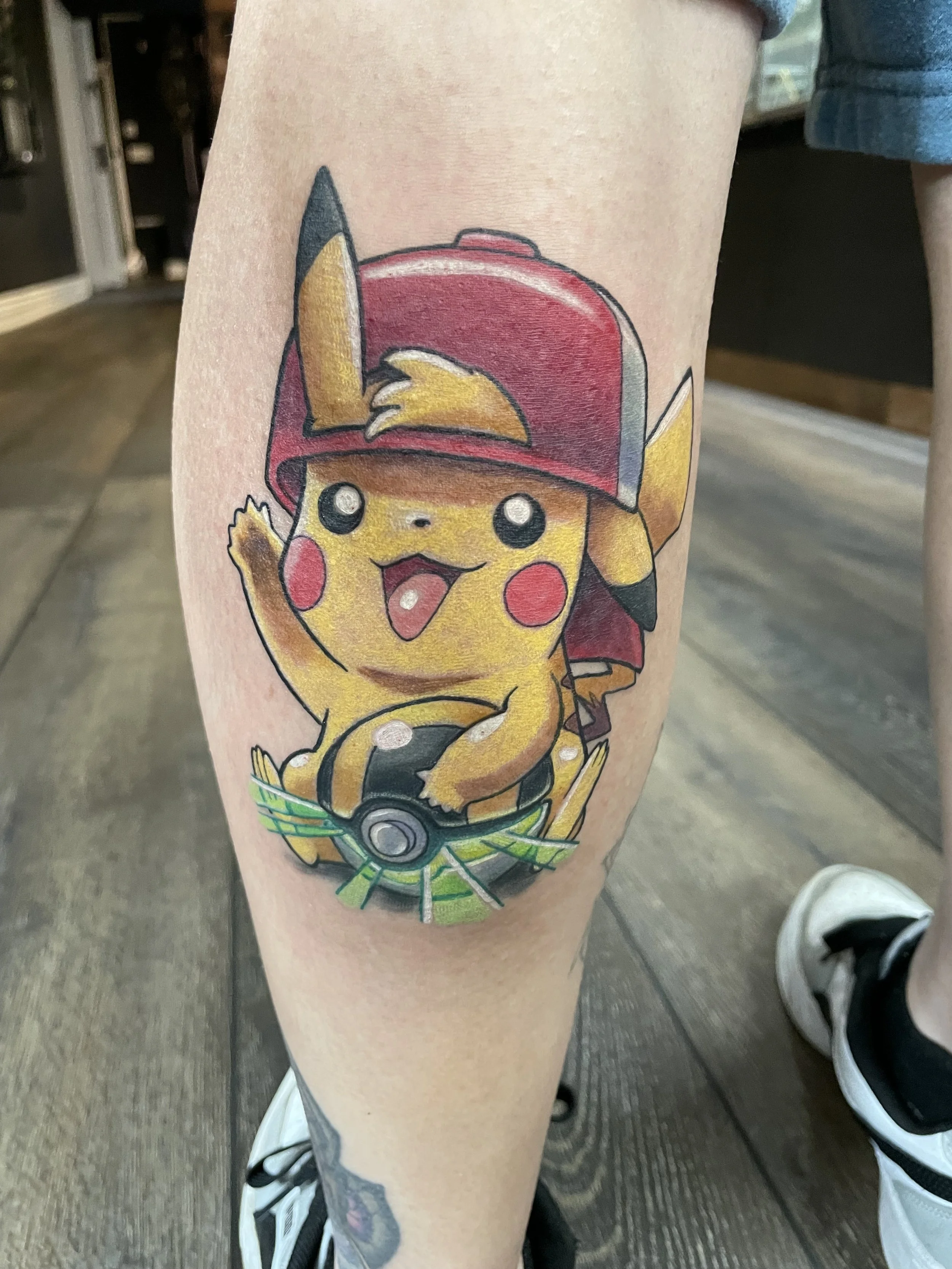 Tattoo of Pikachu from Pokémon, wearing a red cap with a yellow beak and black eyes, in a motorcycle helmet, on a person's thigh.