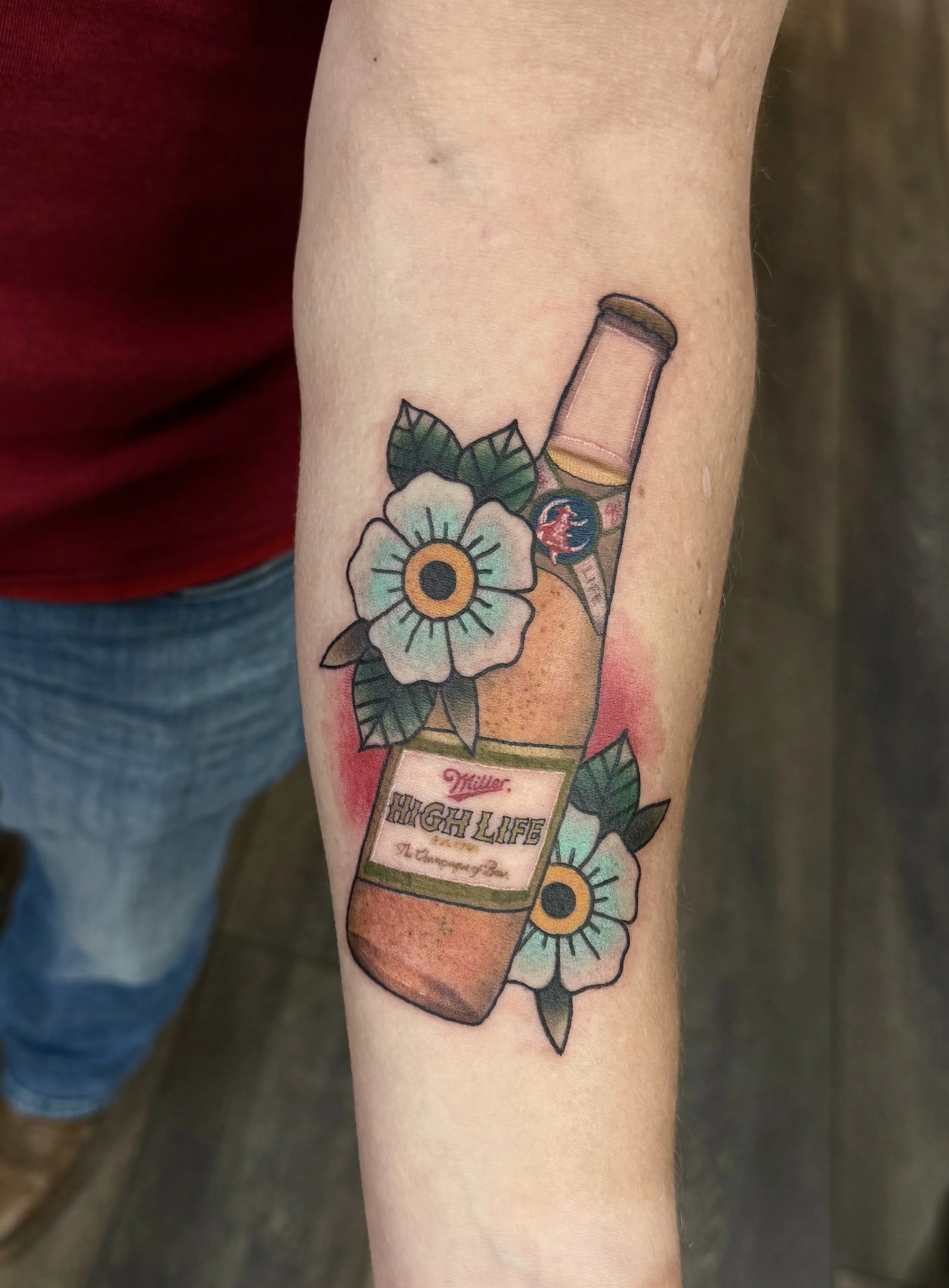 Tattoo of a beer bottle with the label "High Life" and the words "Miller" and "Champagne of Beers", surrounded by a white flower with a yellow center and green leaves, on person's forearm.