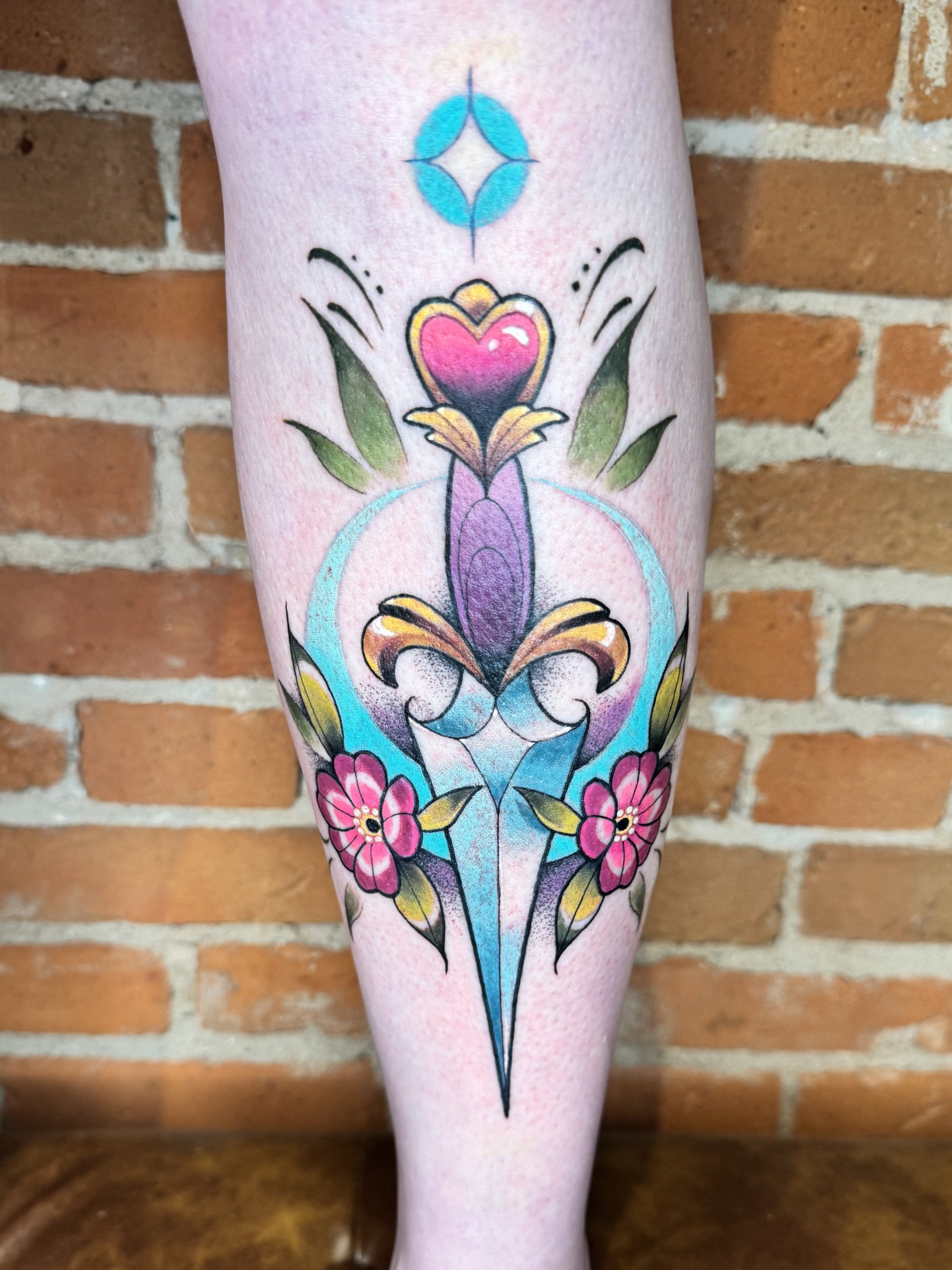 Colorful tattoo on person's arm featuring a central heart shape, floral and ornamental elements, with a blue geometric shape above.