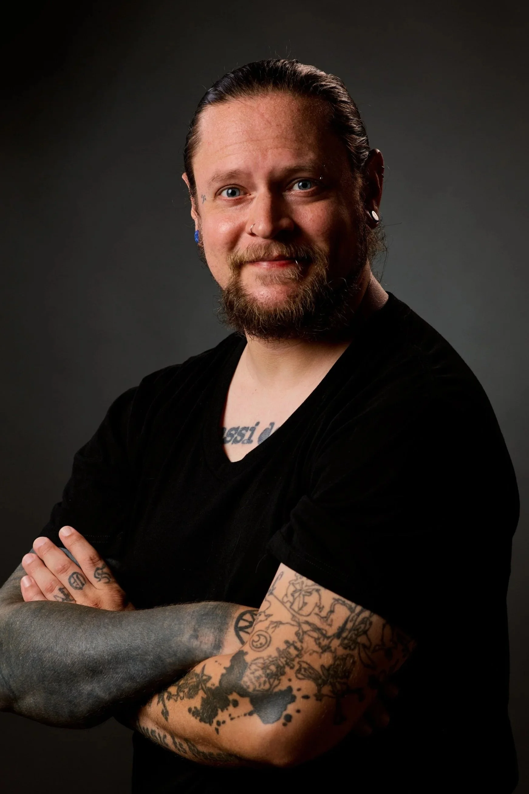 Portrait of a man with tattoos on arms, beard, and piercings, wearing a black shirt, crossing arms, against a dark background.